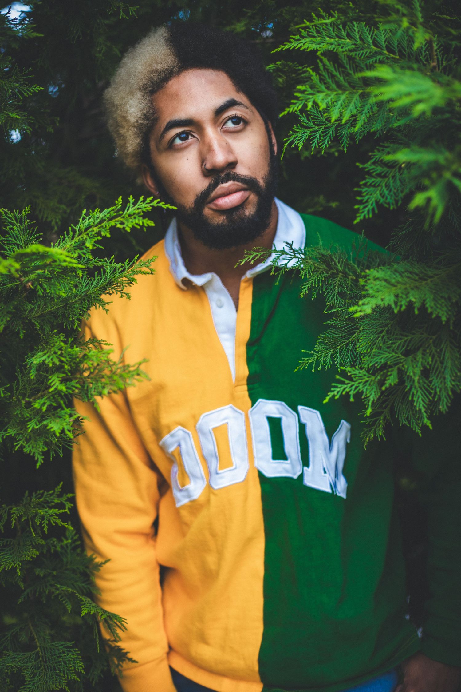Man with afro wearing yellow and green DOOM rugby shirt posing among evergreen trees.