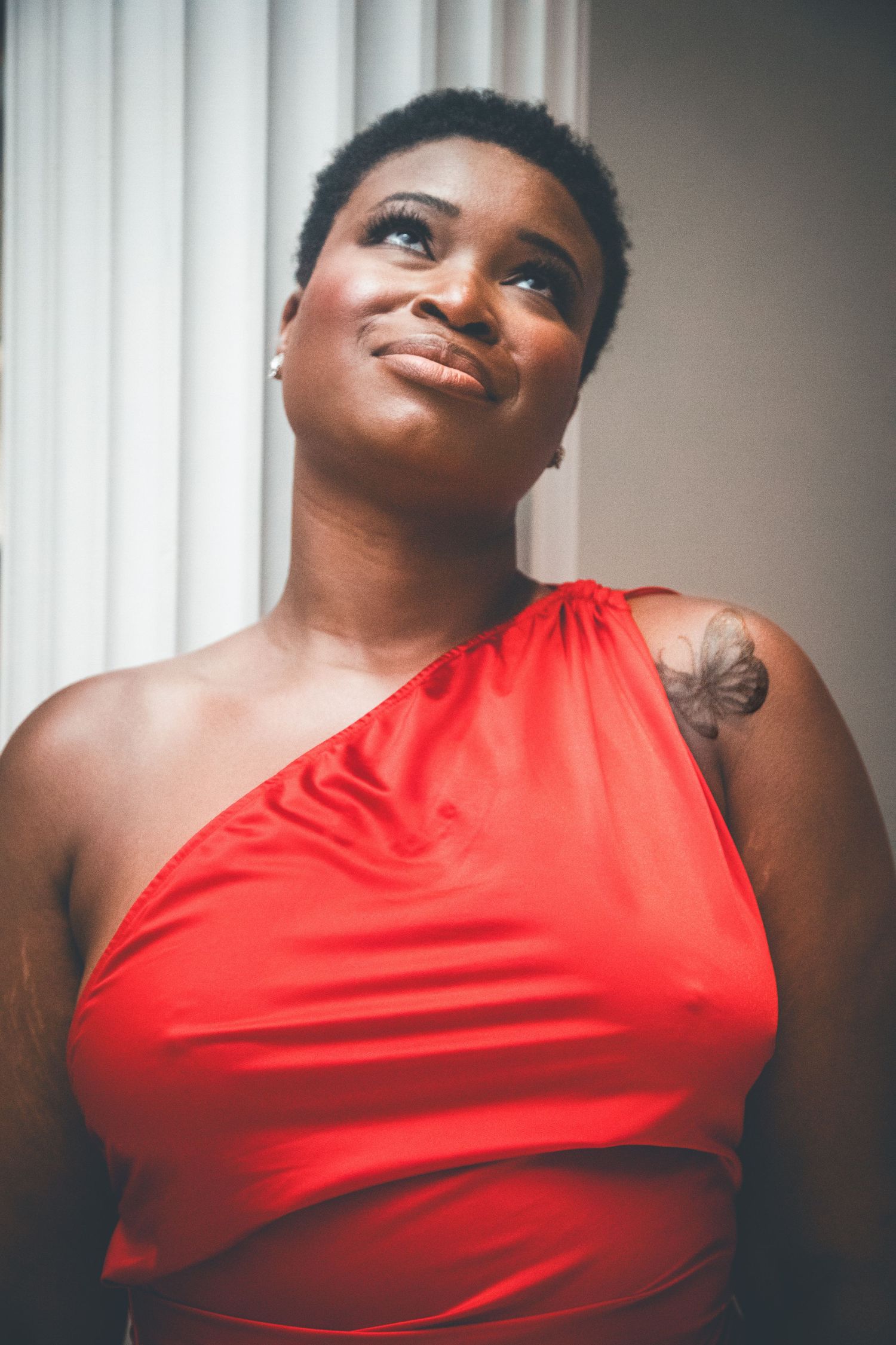 A Black woman in a red one-shoulder dress smiles upward, wearing pearl earrings and a shoulder tattoo.
