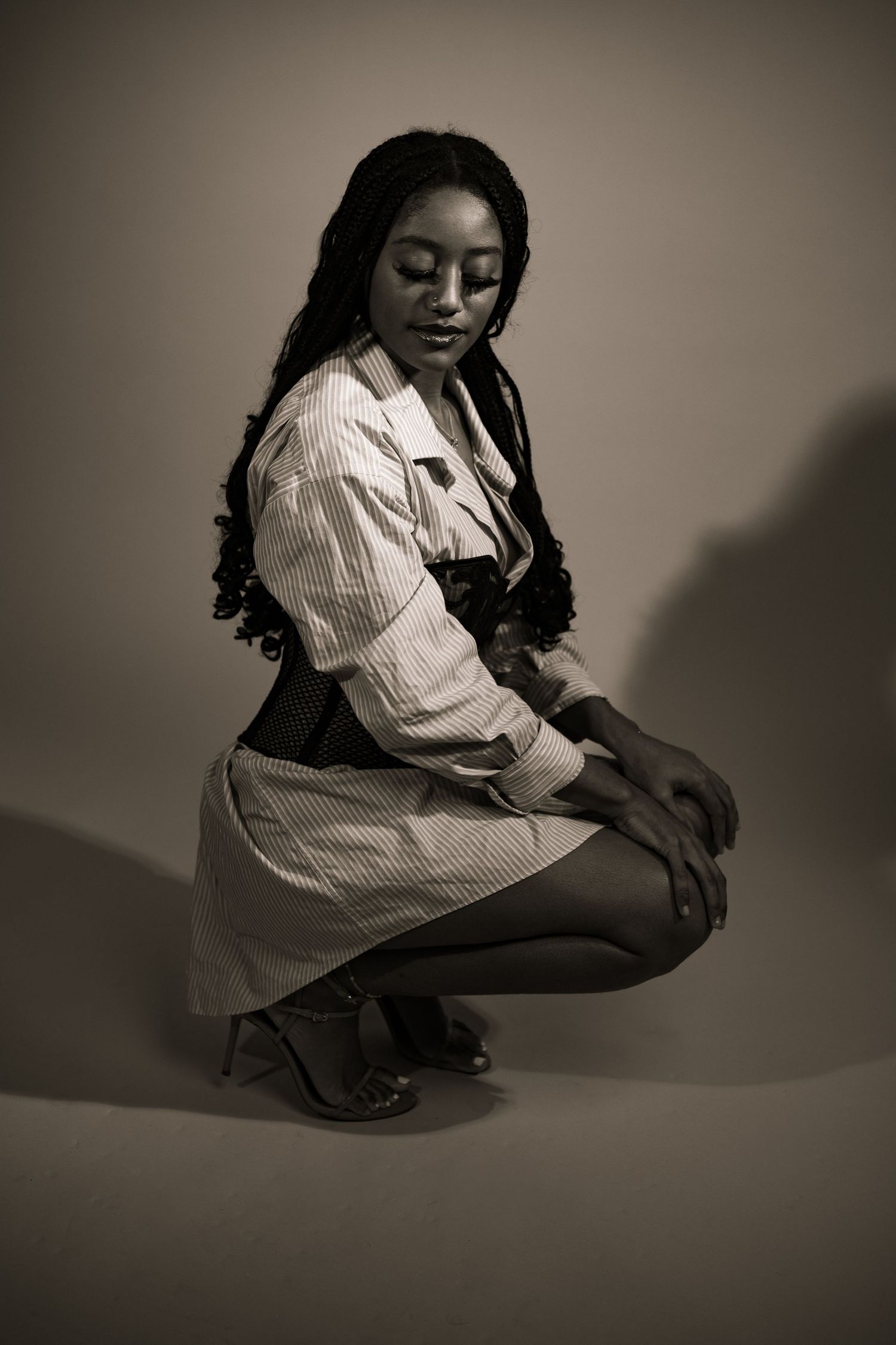 Sepia-toned portrait of a woman with long braids crouching gracefully in a striped outfit and heels.