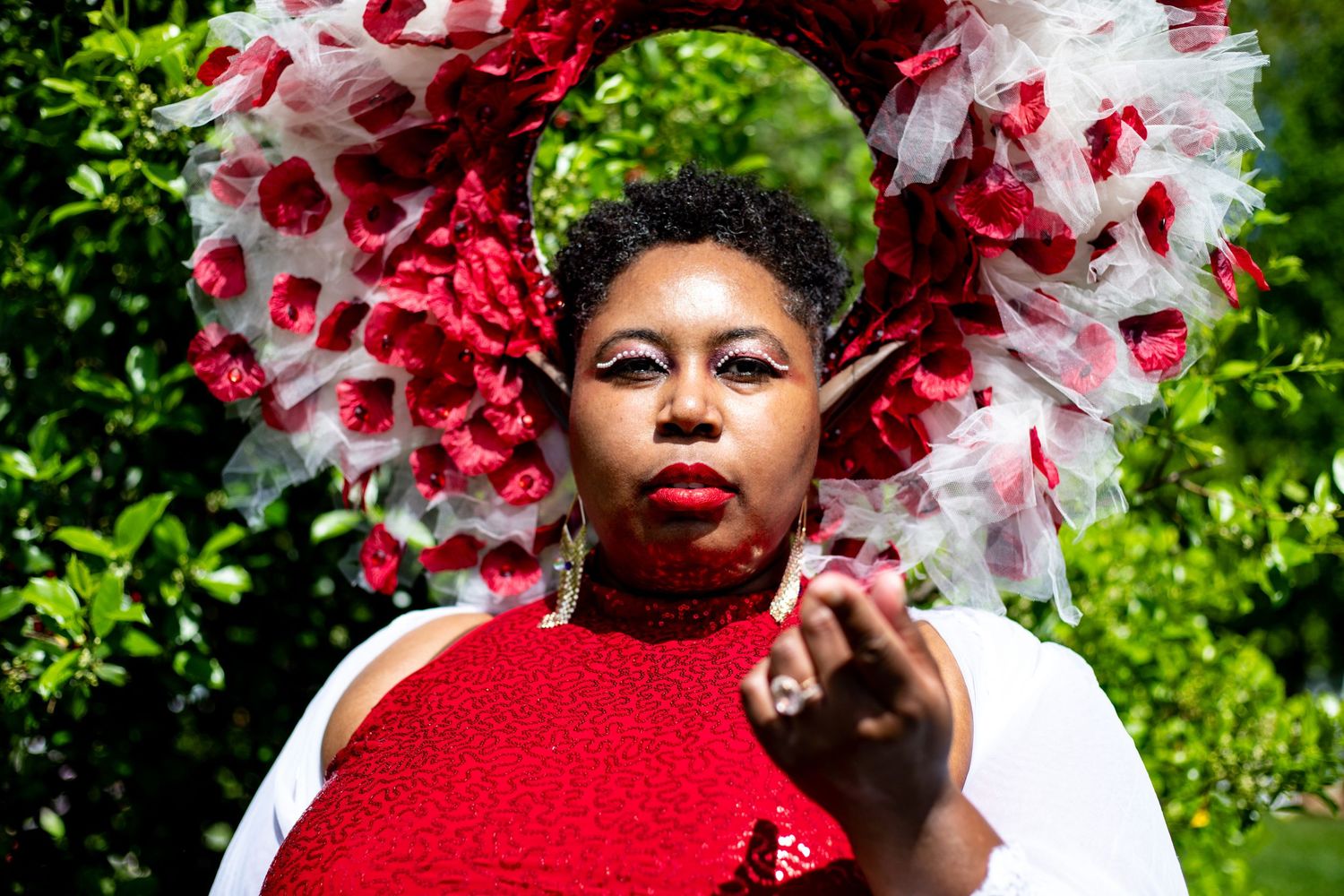 Woman in dramatic red and white floral headpiece and red sequin top poses in garden setting.