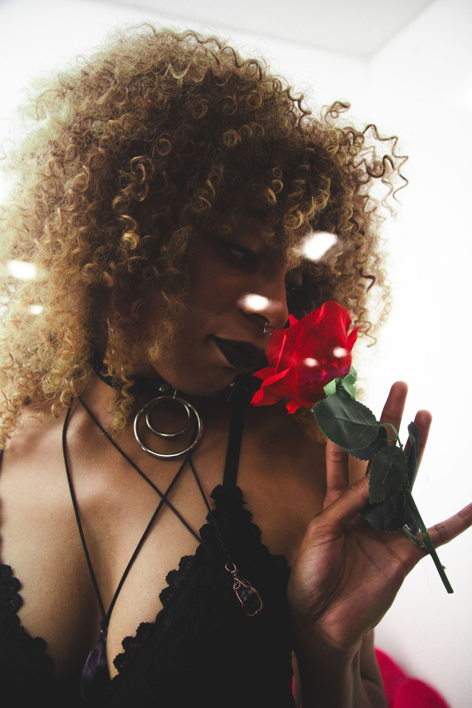 Woman with curly blonde hair holding a red rose near her face, wearing black lace top and choker.