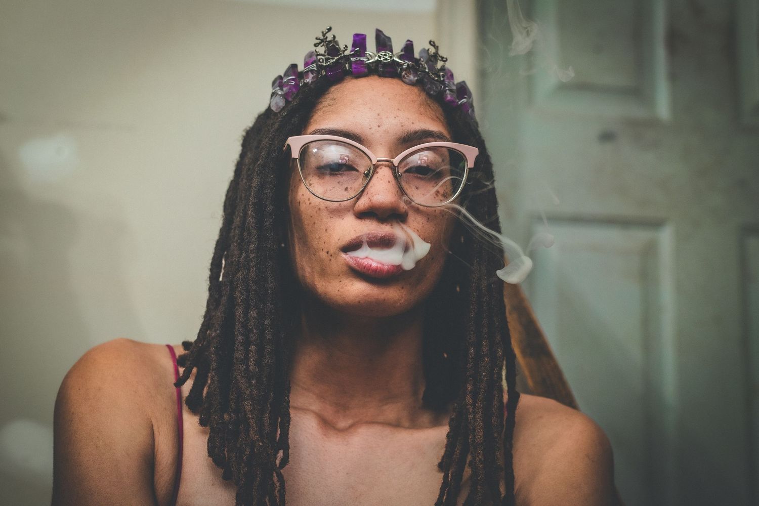 Woman with dreadlocks and floral crown blowing smoke wearing cat-eye glasses.