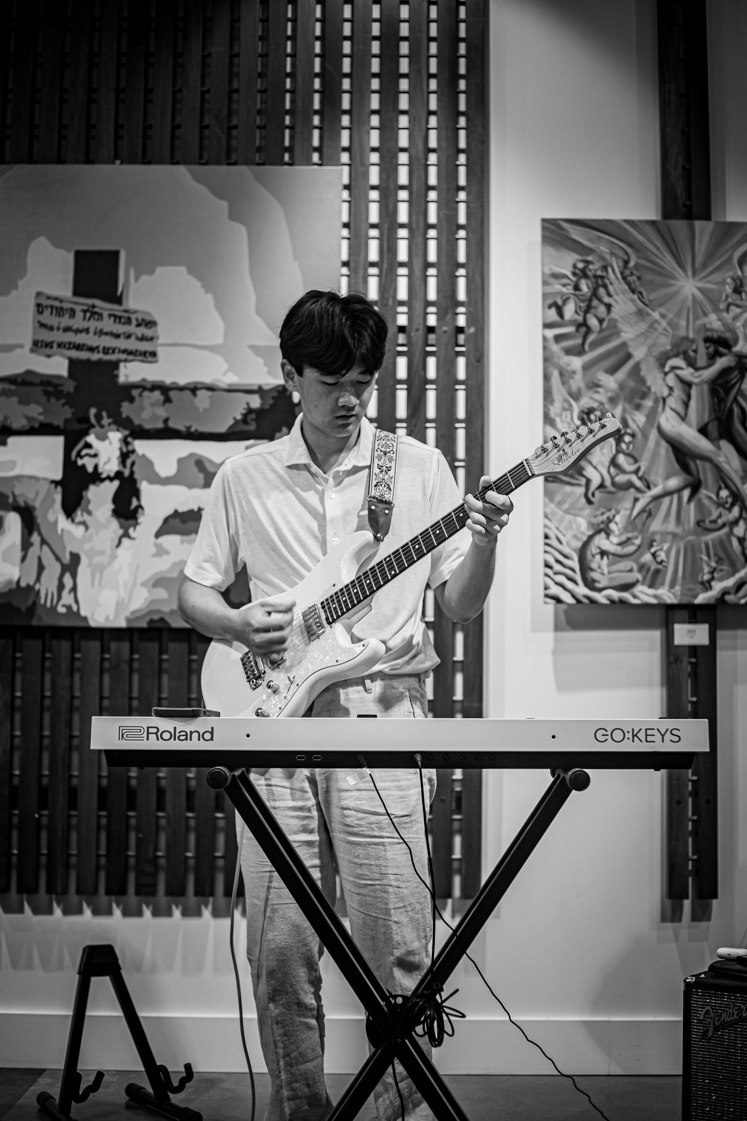 Black and white image of a young man playing electric guitar at an art gallery with paintings in background.