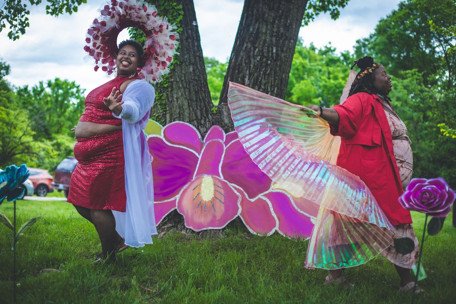 Two women in vibrant red costumes with floral props and iridescent wings pose outdoors at a colorful garden performance.