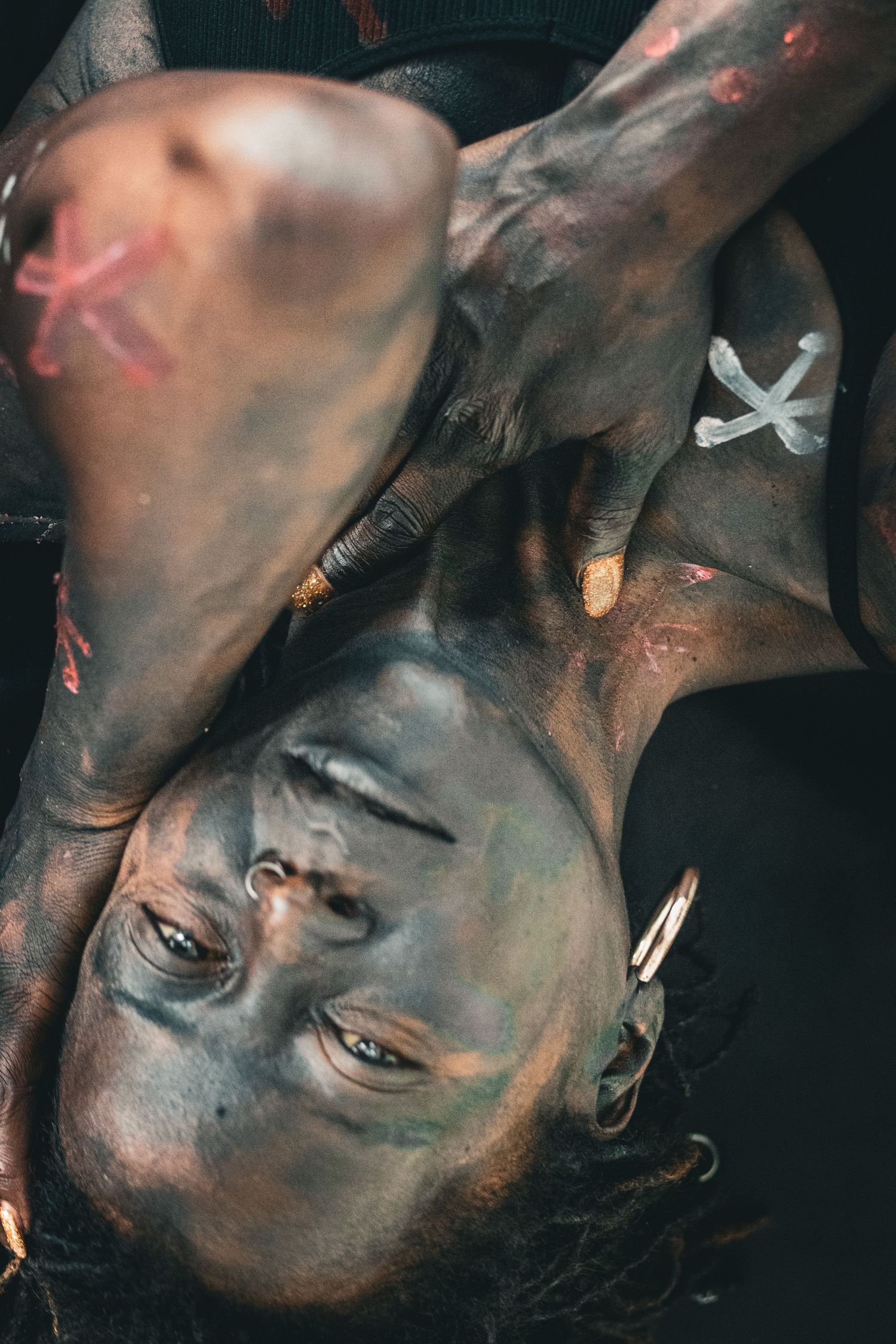 Person with dark body paint and colorful X markings poses upside down in artistic cultural body art photography.