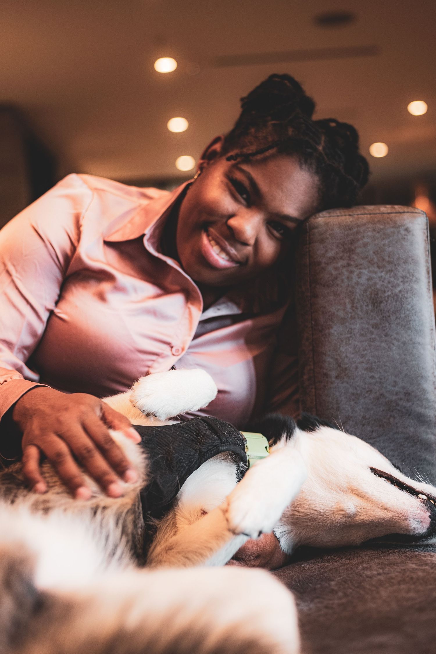 A smiling woman leans on a couch petting a black and white husky lying beside her indoors.