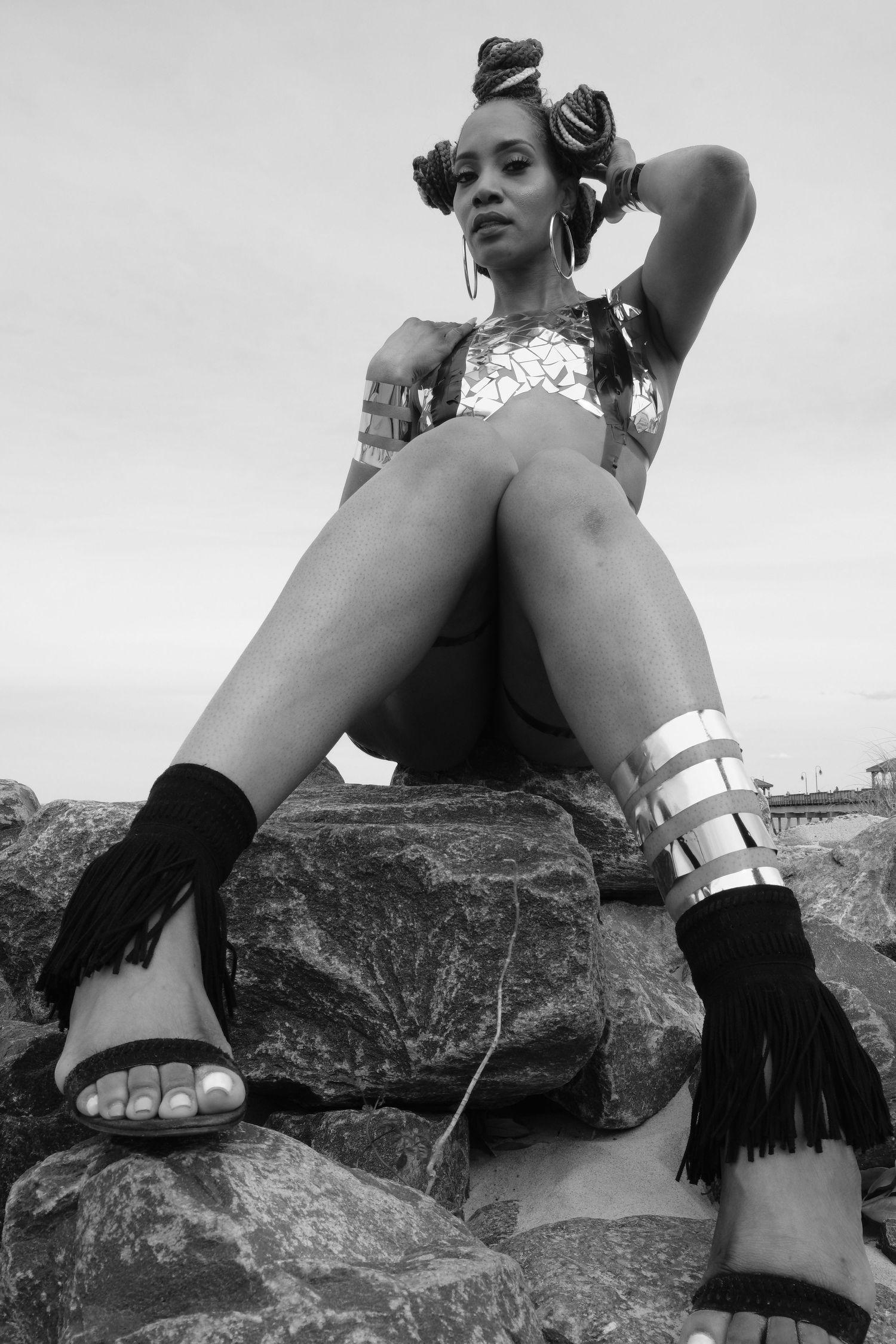 Black and white photo of a woman in festival attire sitting on rocks wearing fringe sandals and metallic accessories.