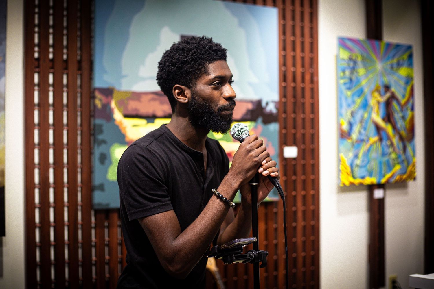 Young Black man singing into a microphone at a colorful art gallery event with vibrant paintings behind him.