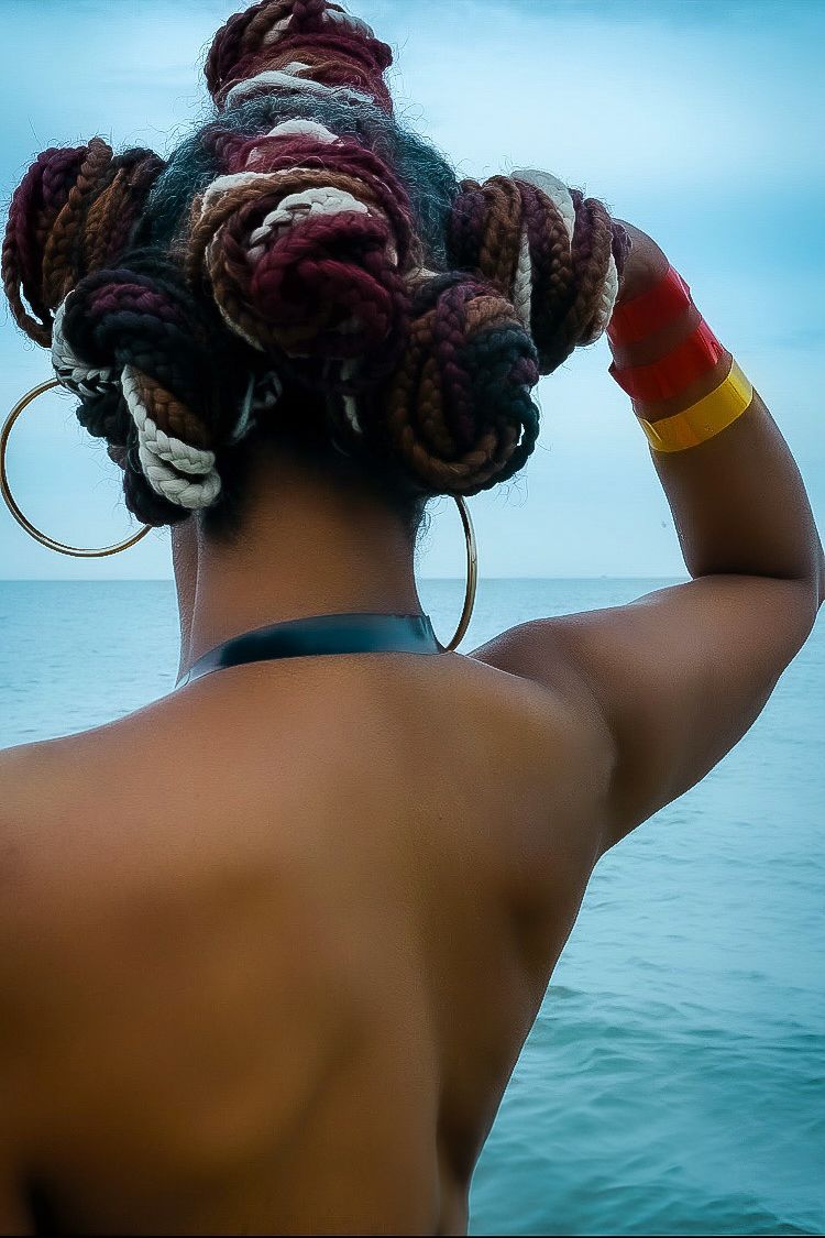 Back view of a woman with colorful braided updo hairstyle wearing hoop earrings by the ocean.