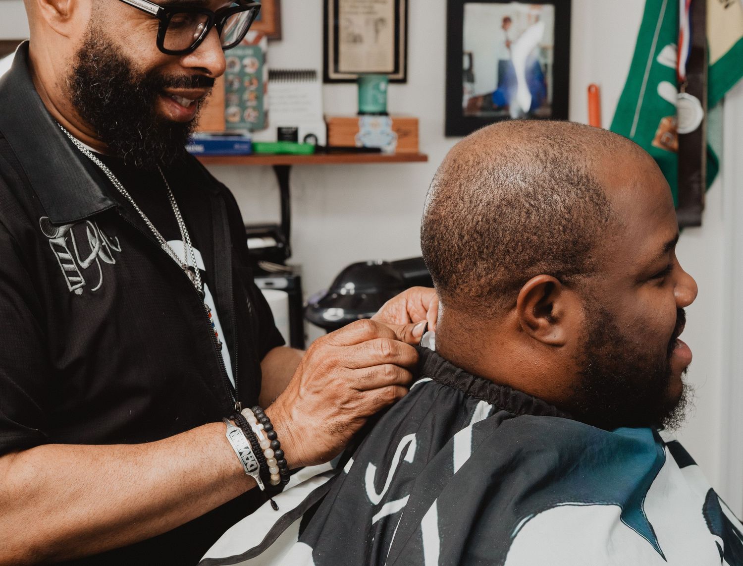 Smiling barber with glasses trimming a bearded client in a professional barbershop.
