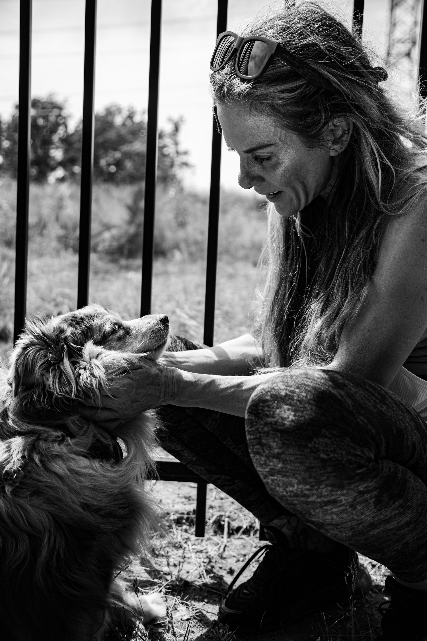 Black and white photo of a smiling woman with sunglasses petting a small dog near metal fence outdoors.