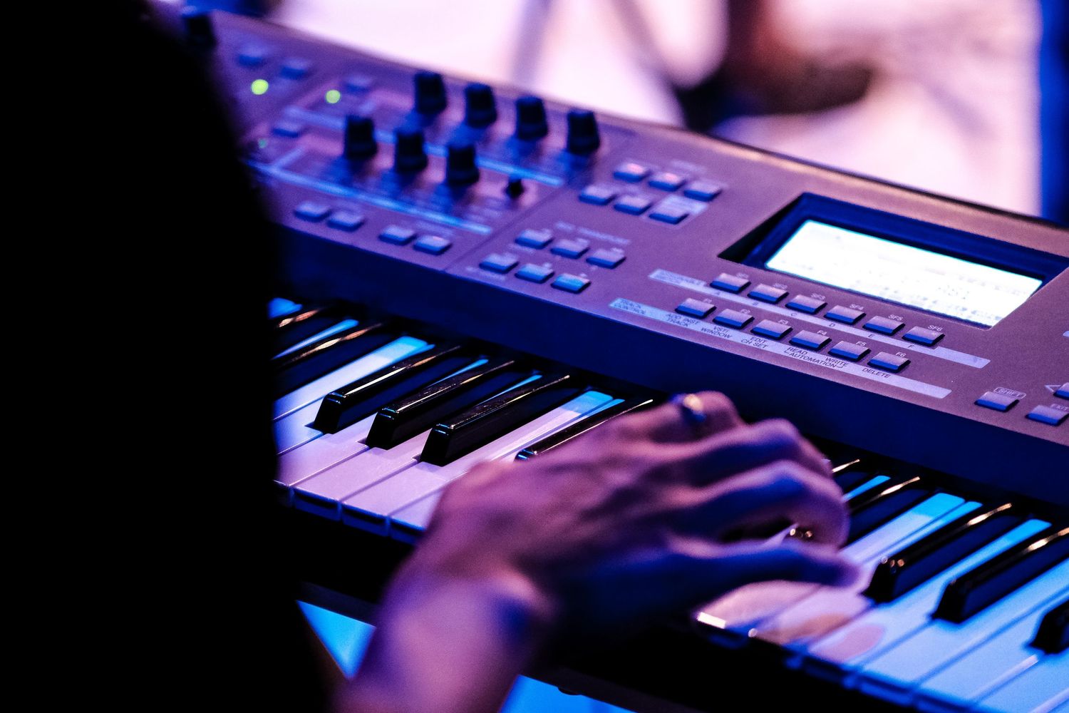 Musician playing a professional keyboard synthesizer under blue and purple stage lighting.