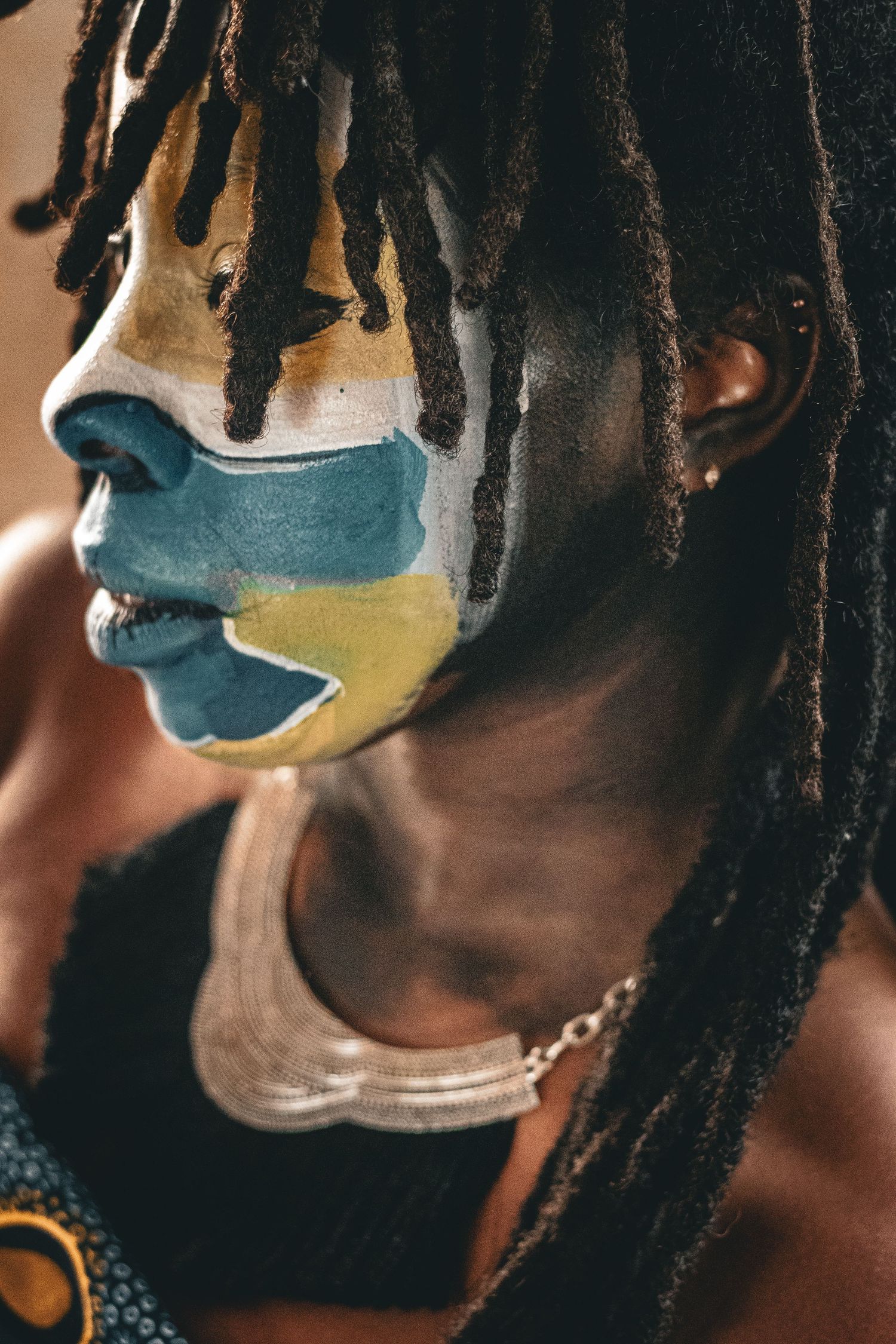 Profile portrait of person with dreadlocks and vibrant blue and yellow face paint artistic makeup.