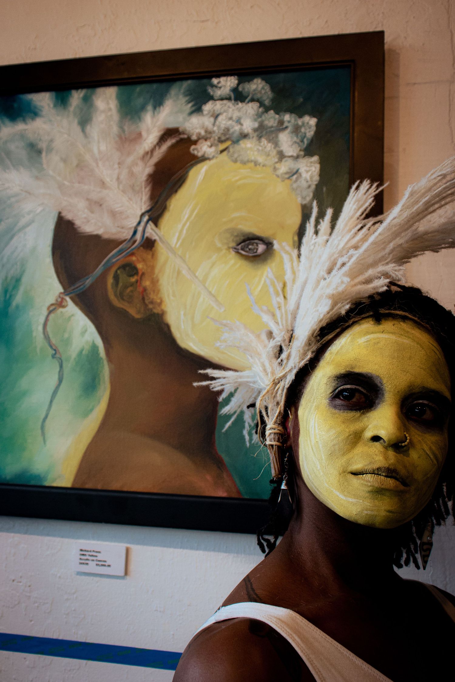 Woman with yellow face paint stands beside a matching painted portrait with white feathers.