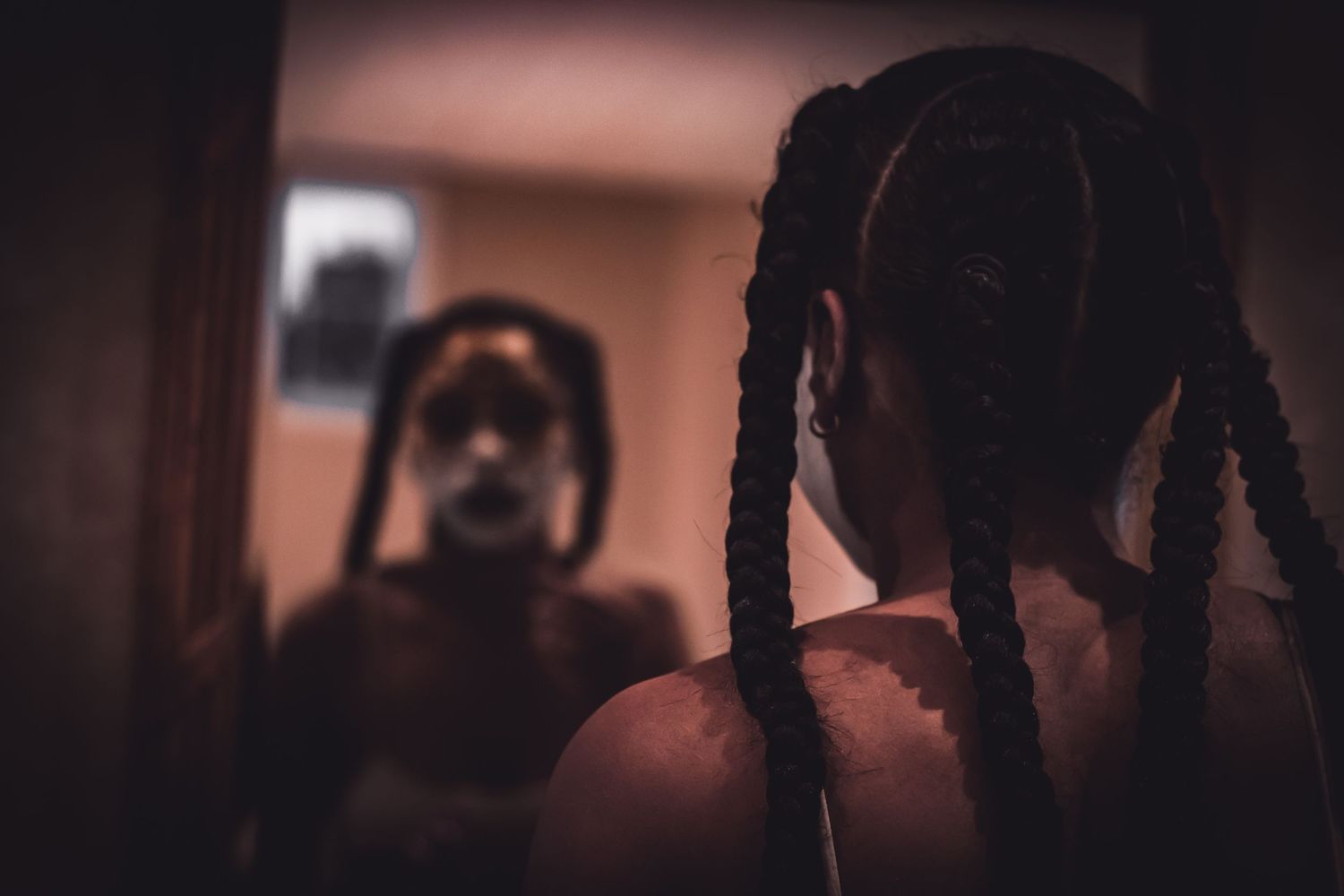 Person with braided hair looking at their reflection in a dimly lit mirror with face paint.
