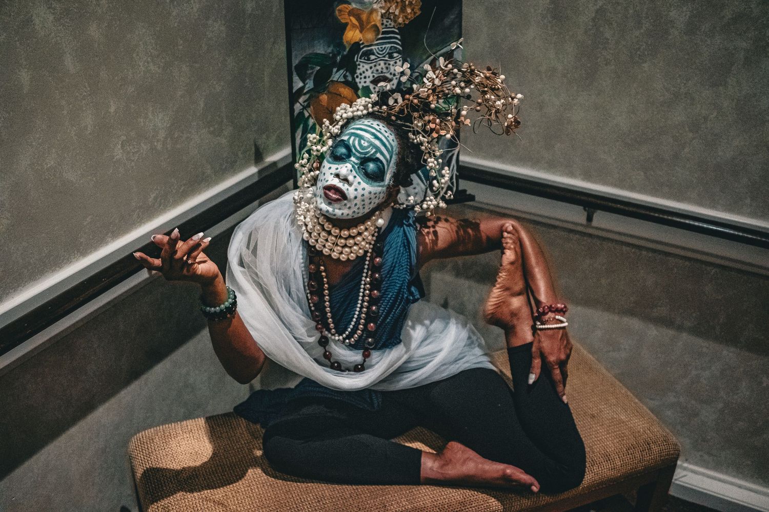 Performer in elaborate blue face paint, floral headdress, pearls, and traditional costume seated on bench.