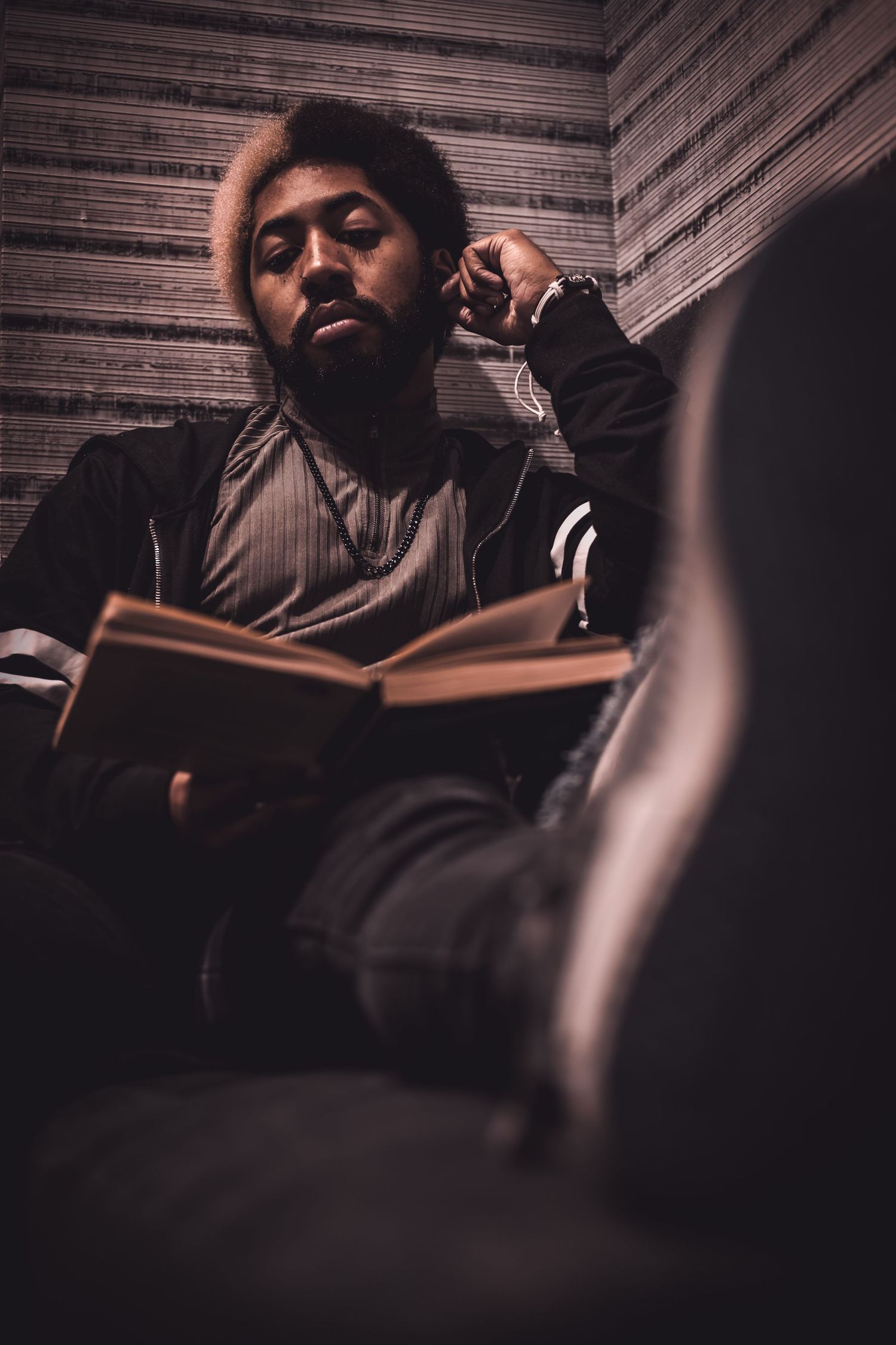 Young Black man with an afro reading a book while listening to music in a dimly lit room.