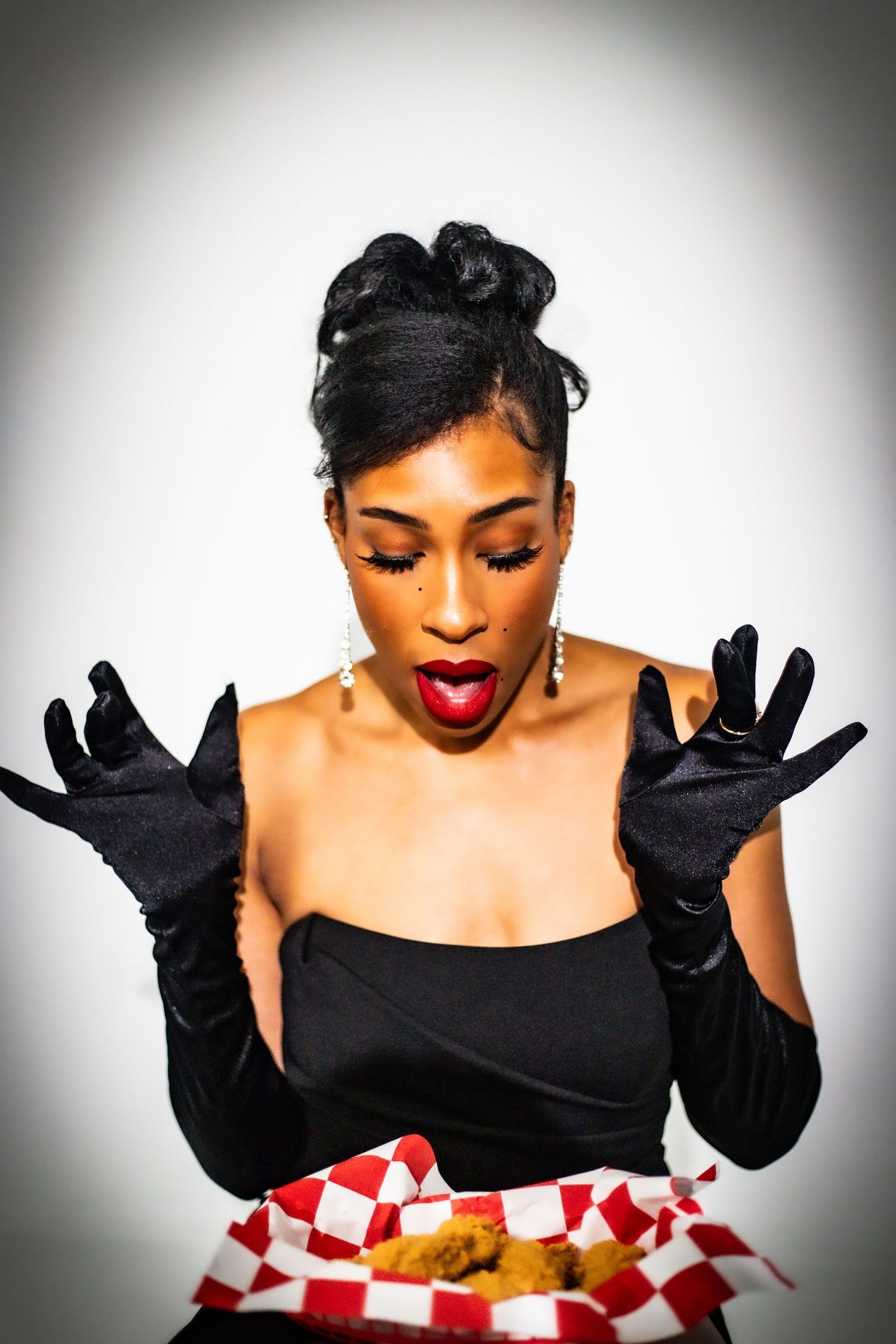 Elegant woman in black strapless dress and long gloves reacting with surprise to a basket of fried chicken.