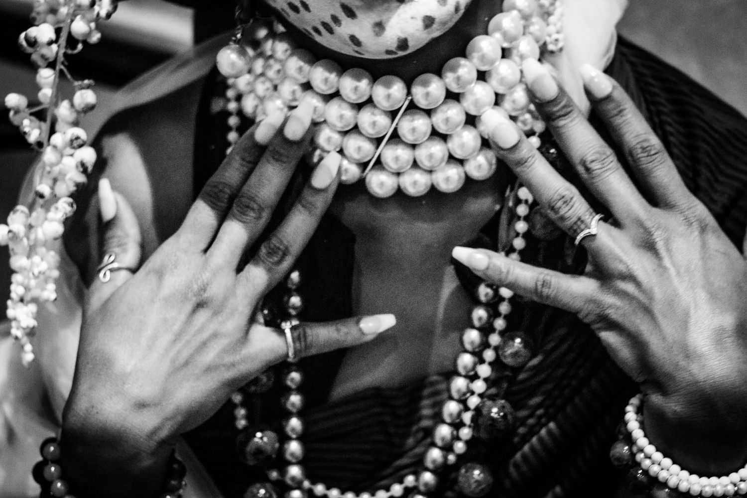 Close-up black and white photo of hands adorned with rings and long nails over pearl necklaces.