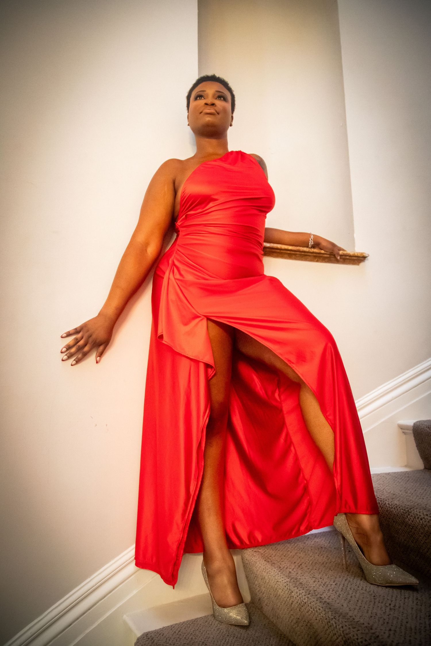 Black woman in a red satin one-shoulder gown with high slit poses on stairs against a white wall.