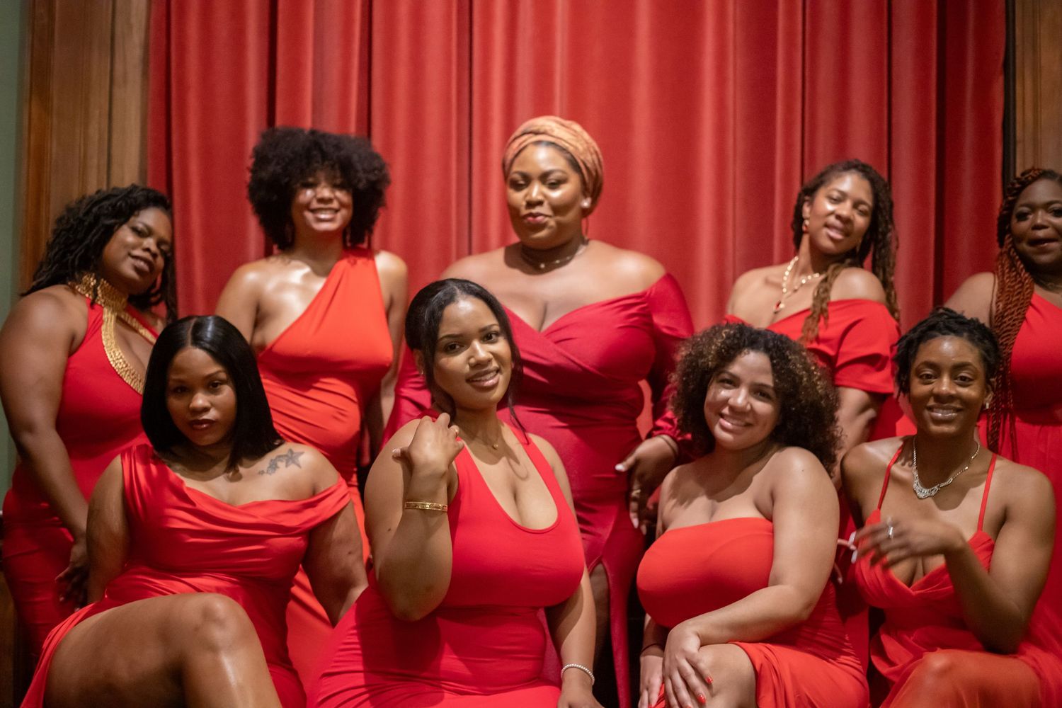 Group of women in matching red dresses posing together for a formal photo against red curtain backdrop.