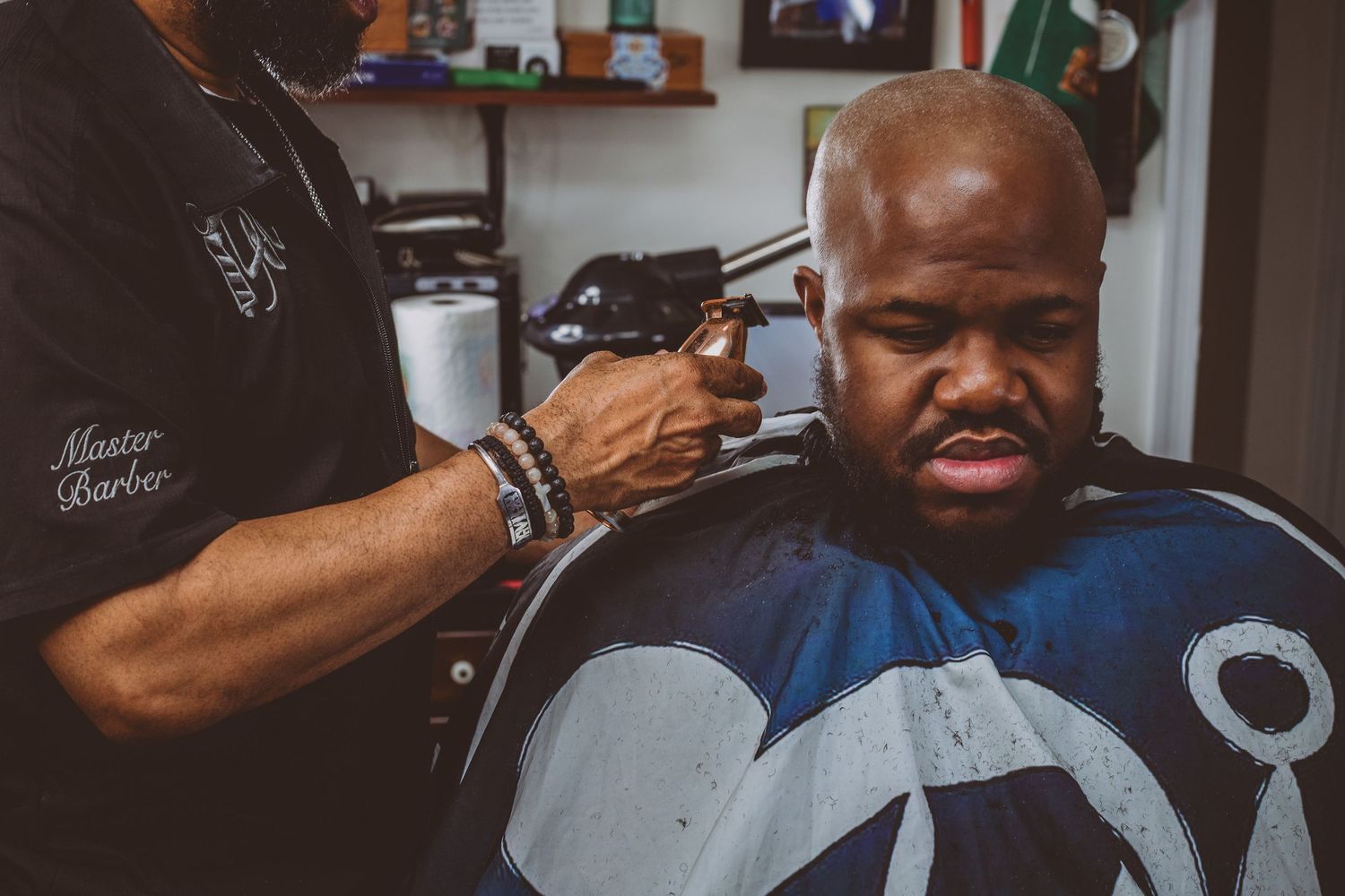 Master barber using clippers on a bald client wearing a blue cape in a barbershop.