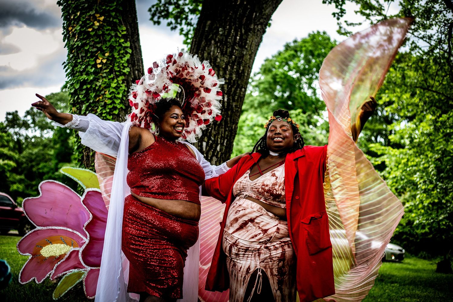 Two joyful women in elaborate red floral costumes pose together outdoors with colorful butterfly wings and flower props.