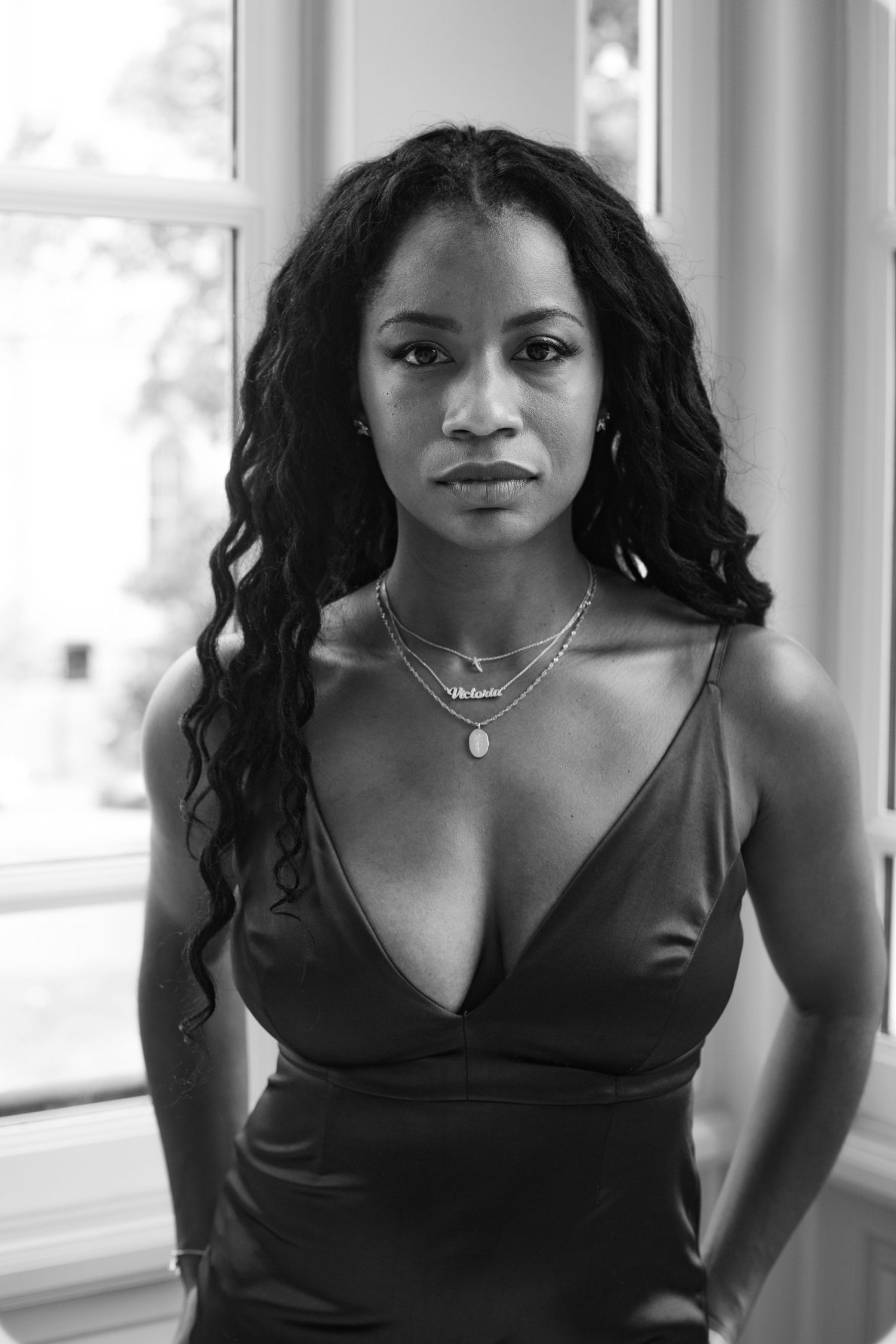 Black and white portrait of Black woman in satin dress with layered necklaces by window.