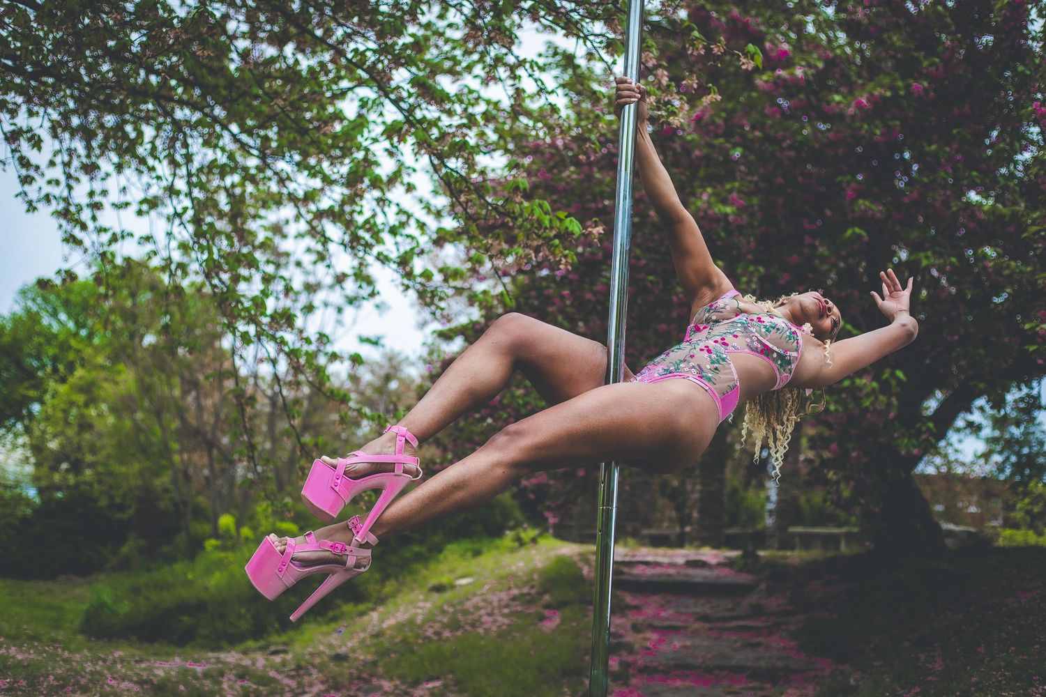 Blonde woman in pink heels performing aerial pole dance outdoors among spring trees.
