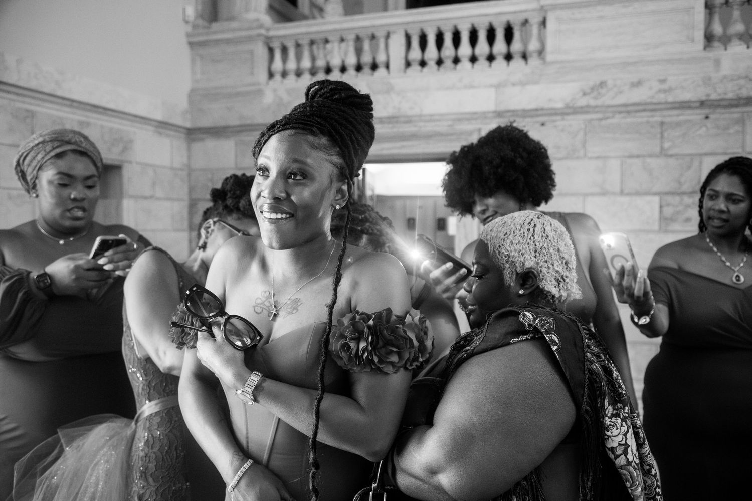 Black and white photo of women helping a smiling bride get ready at an elegant venue.