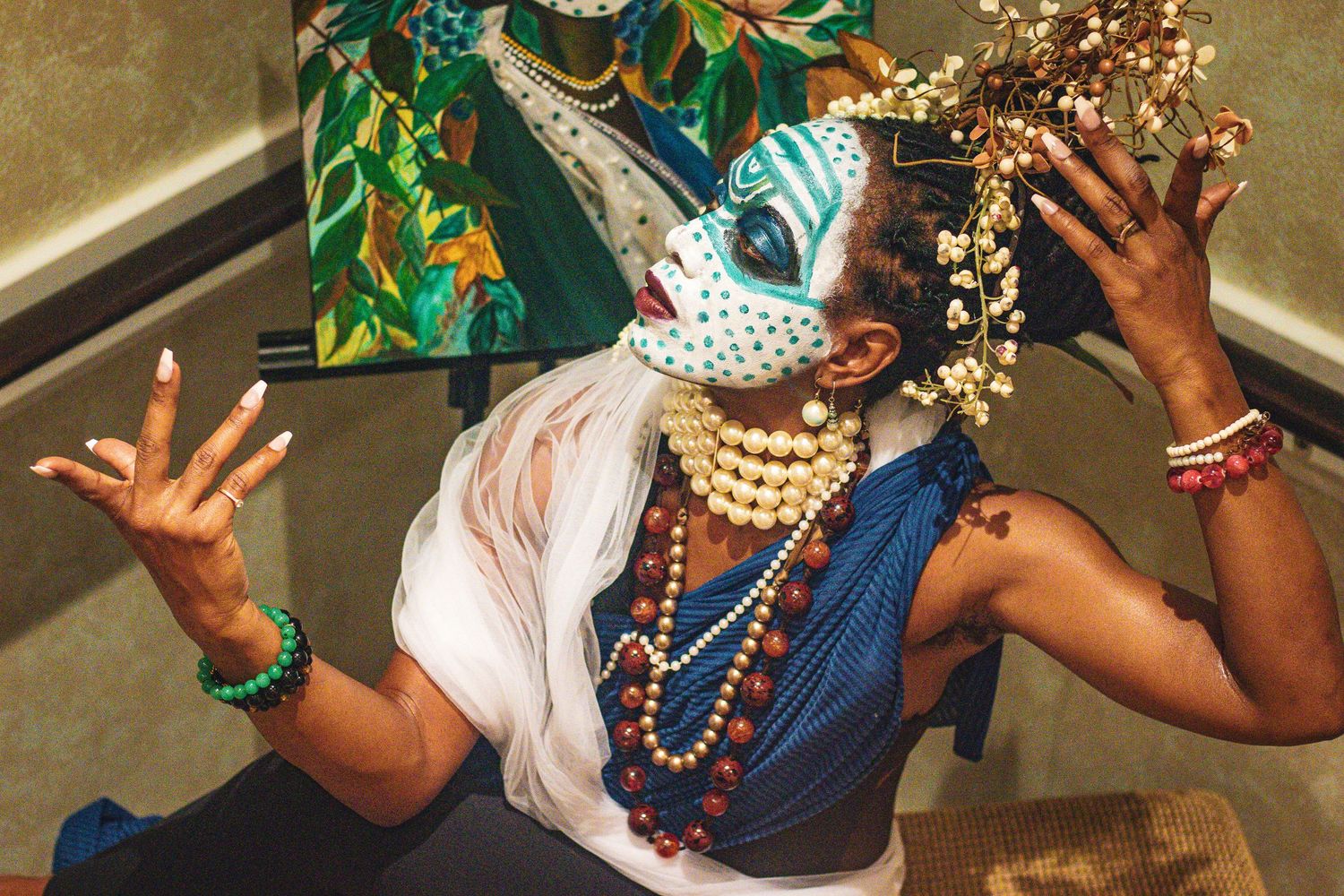 Performer in blue face paint and floral headdress gesturing with hands adorned with pearl necklaces.