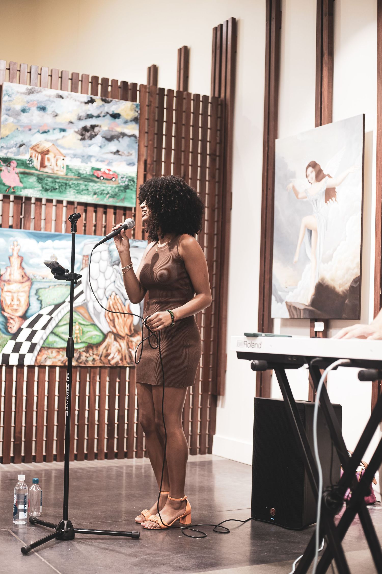 Female performer in brown dress singing at microphone stand surrounded by colorful artwork.