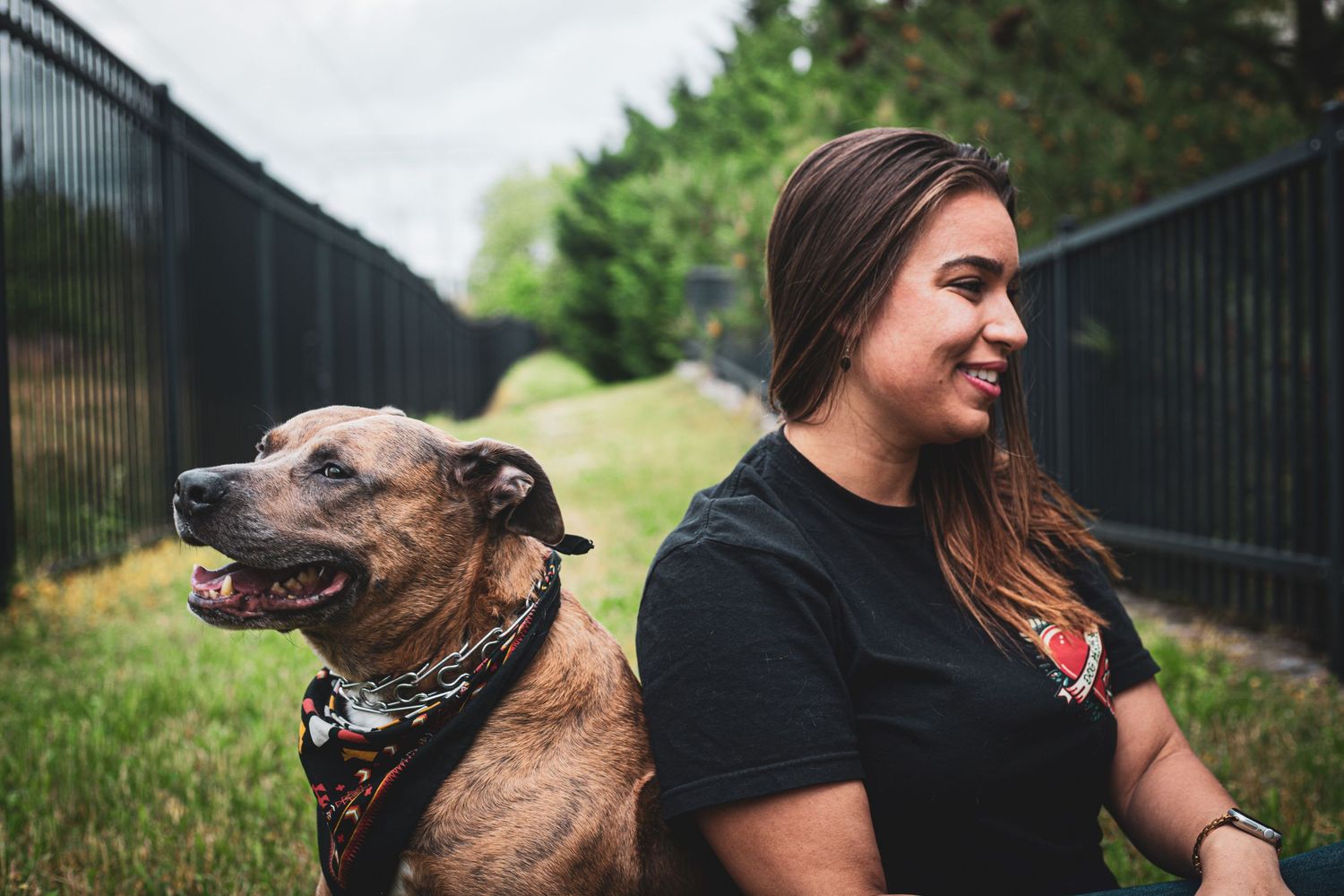 Woman in black tee smiling beside a brindle, pitbull-mix dog wearing a bandana near a black fence outdoors.
