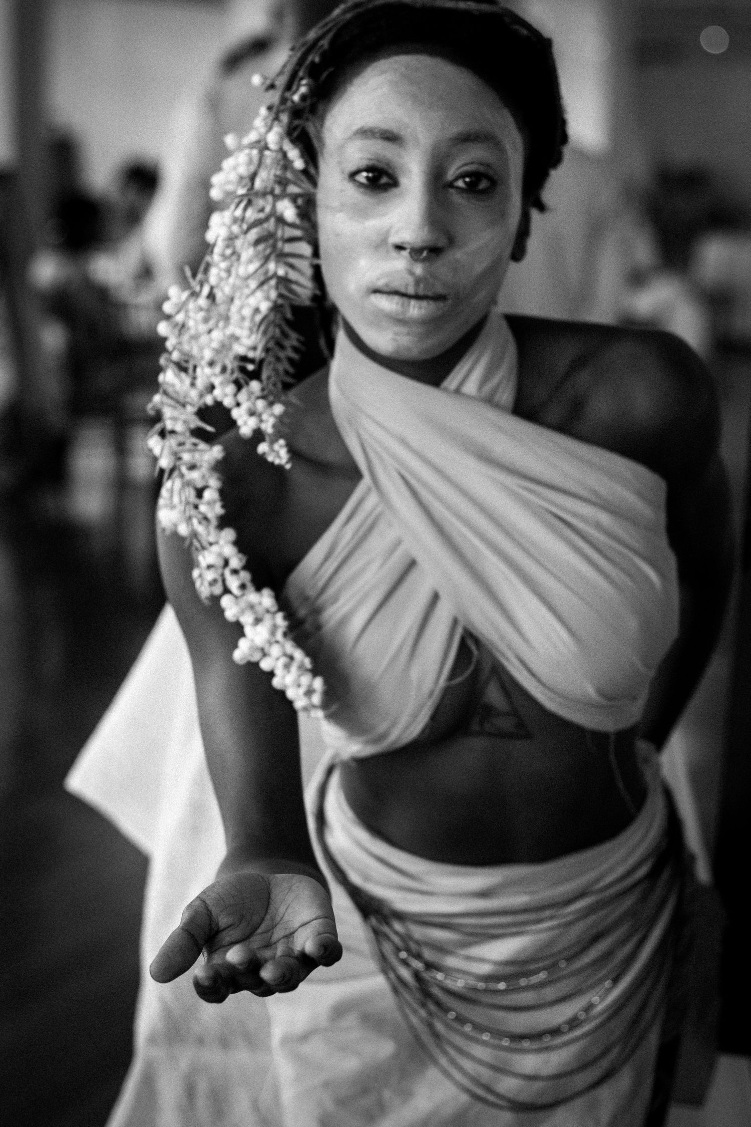Black and white portrait of a model with white face paint in draped fabric costume extending open hand forward.