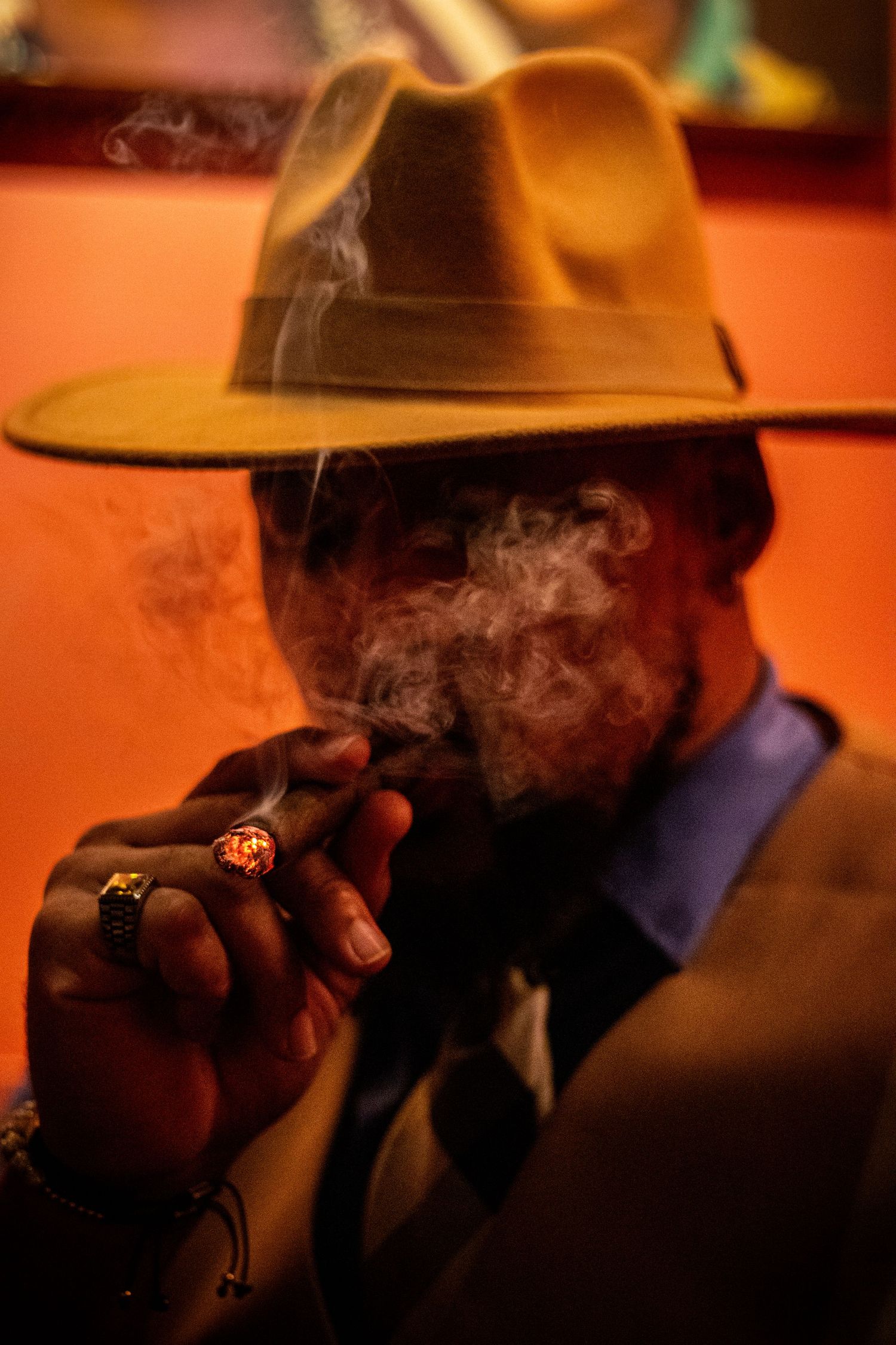 Stylish man in fedora hat smoking a cigar with smoke swirling in warm orange ambient light.