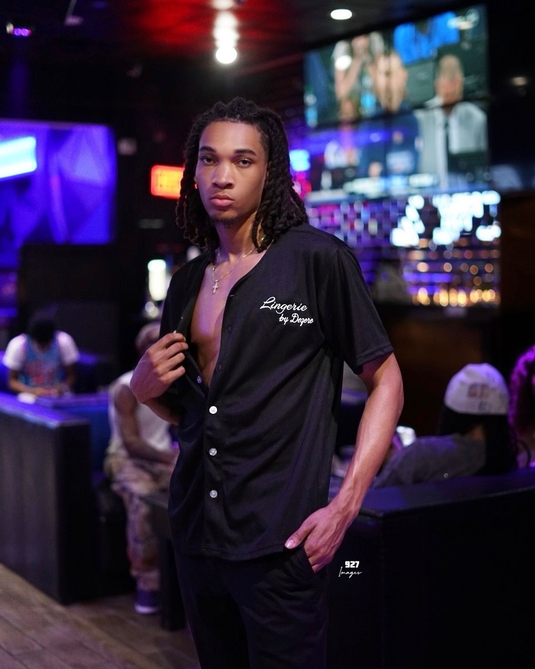 Young man with braids wearing a black button-up shirt poses confidently in a vibrant nightclub setting.