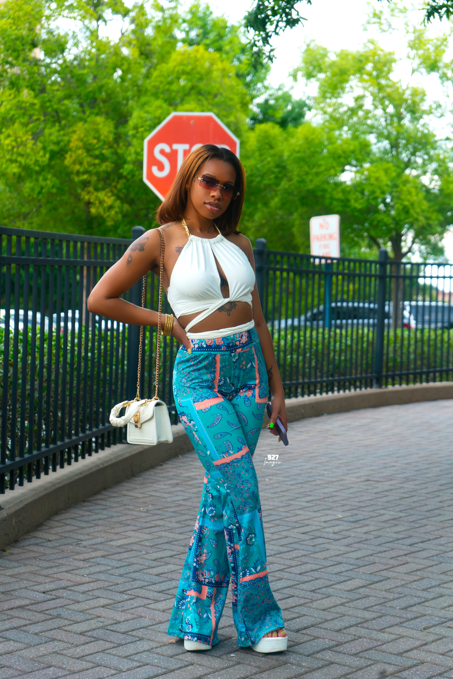 A stylish woman poses outdoors in a white crop top and blue floral flare pants with a white bag near a stop sign.