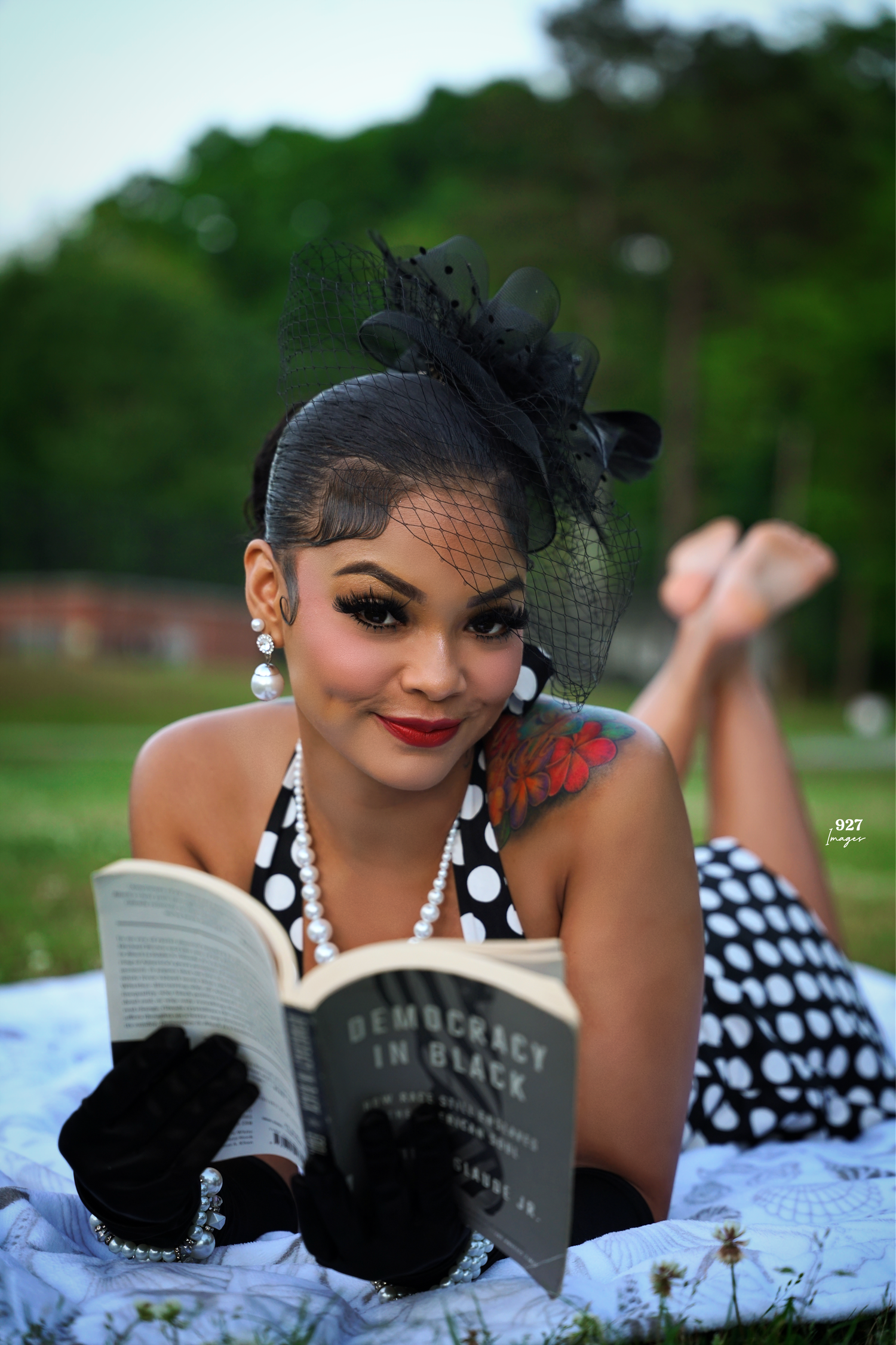 Elegant woman in polka dot dress reading Democracy in Black book while posing outdoors with fascinator hat.