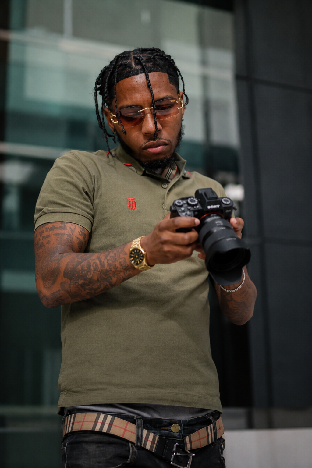 Tattooed man in olive polo shirt reviewing photos on Sony DSLR camera outdoors near glass building.