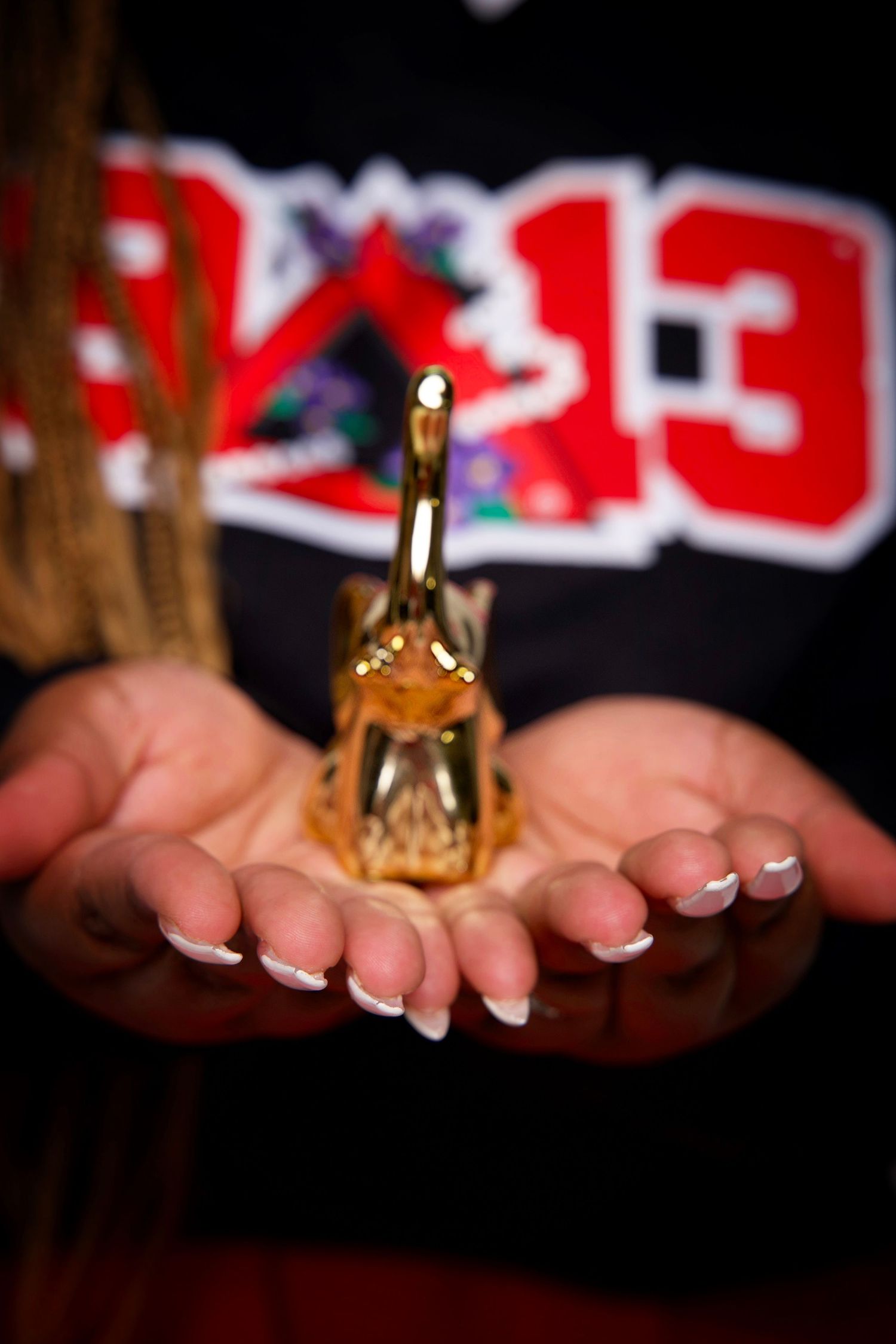 Person holding a small golden trophy in outstretched palms, wearing a black and red varsity jacket with French manicured nails.