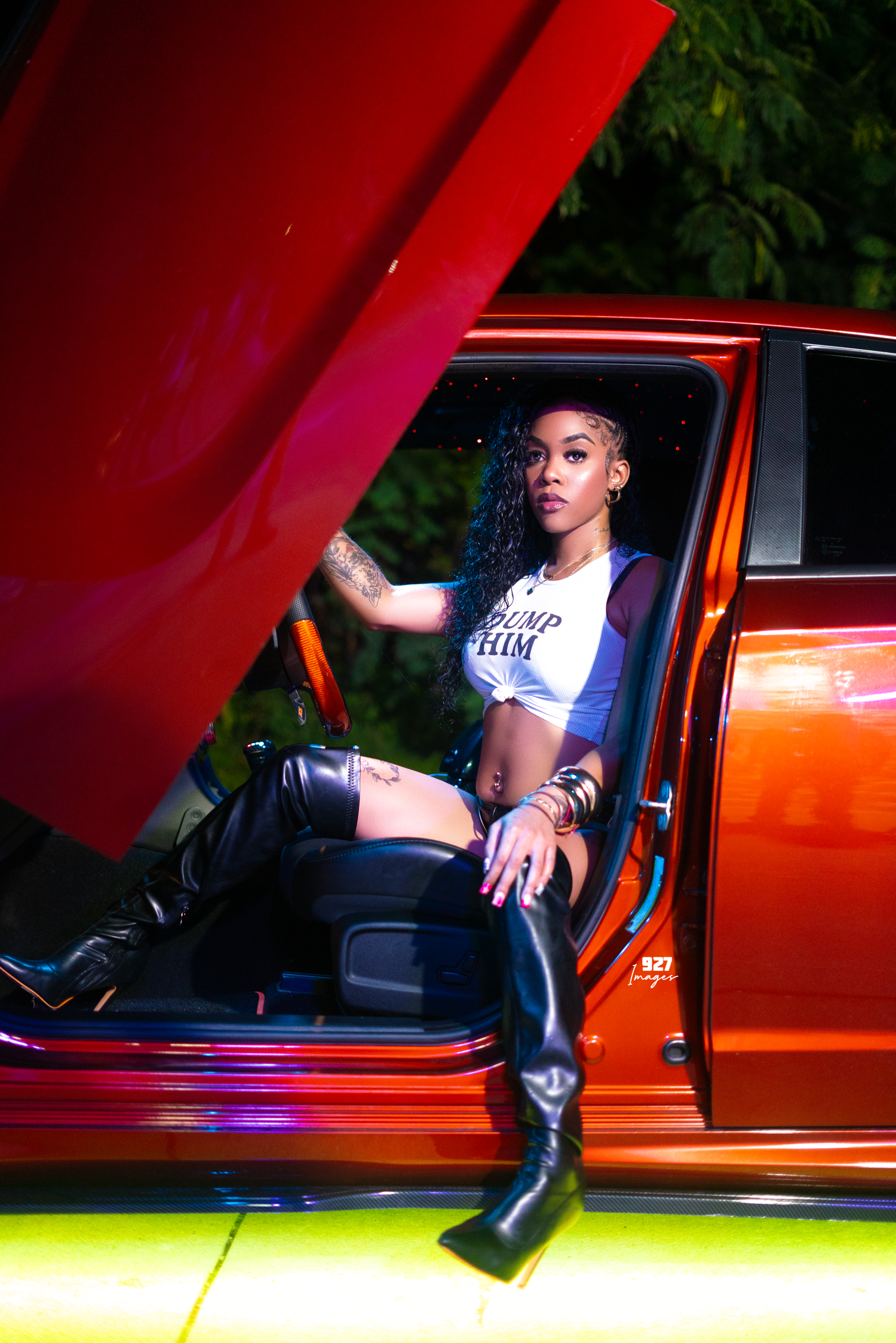 Woman in thigh-high boots posing inside a custom orange lowrider car with hood open at night.