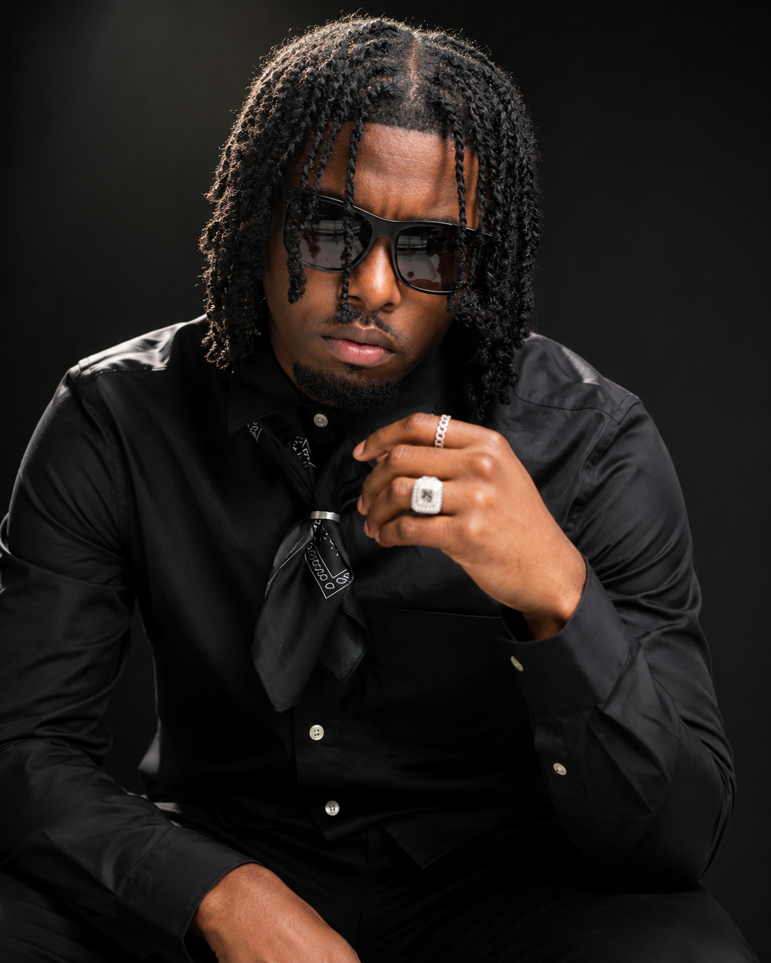 Stylish young man in all-black attire and sunglasses posing with diamond ring in dramatic studio lighting.