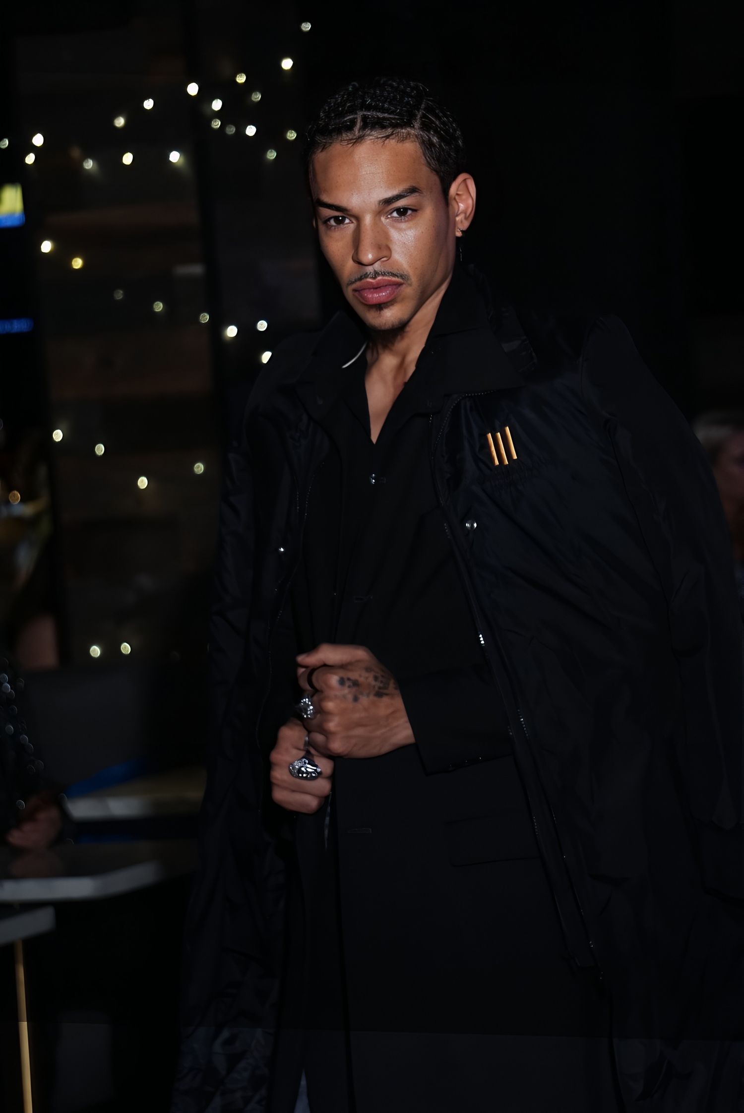 A young man in an all-black outfit and braided hair poses confidently against a dark bokeh background.