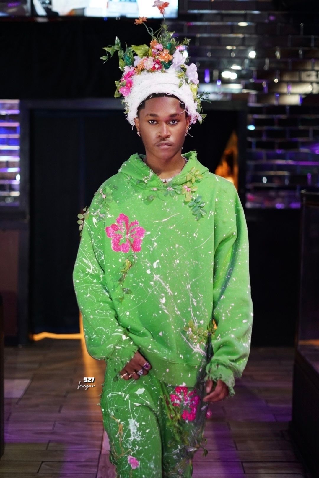 Model wearing green floral hoodie set with paint splatter and floral headpiece on fashion runway.