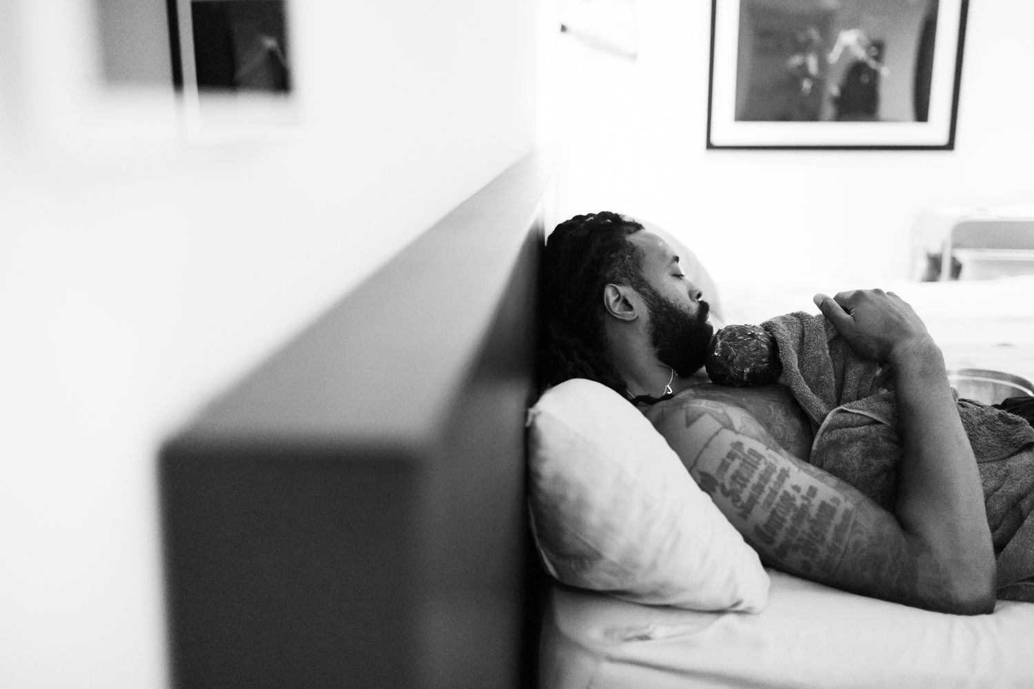 Black and white photo of a tattooed man resting on a bed, leaning against the headboard in a relaxed pose.