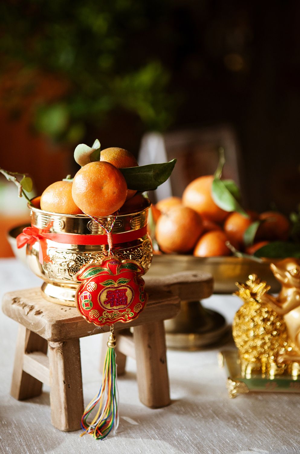 A golden bowl of fresh mandarin oranges with a red Chinese lucky charm ornament on a wooden stool display.