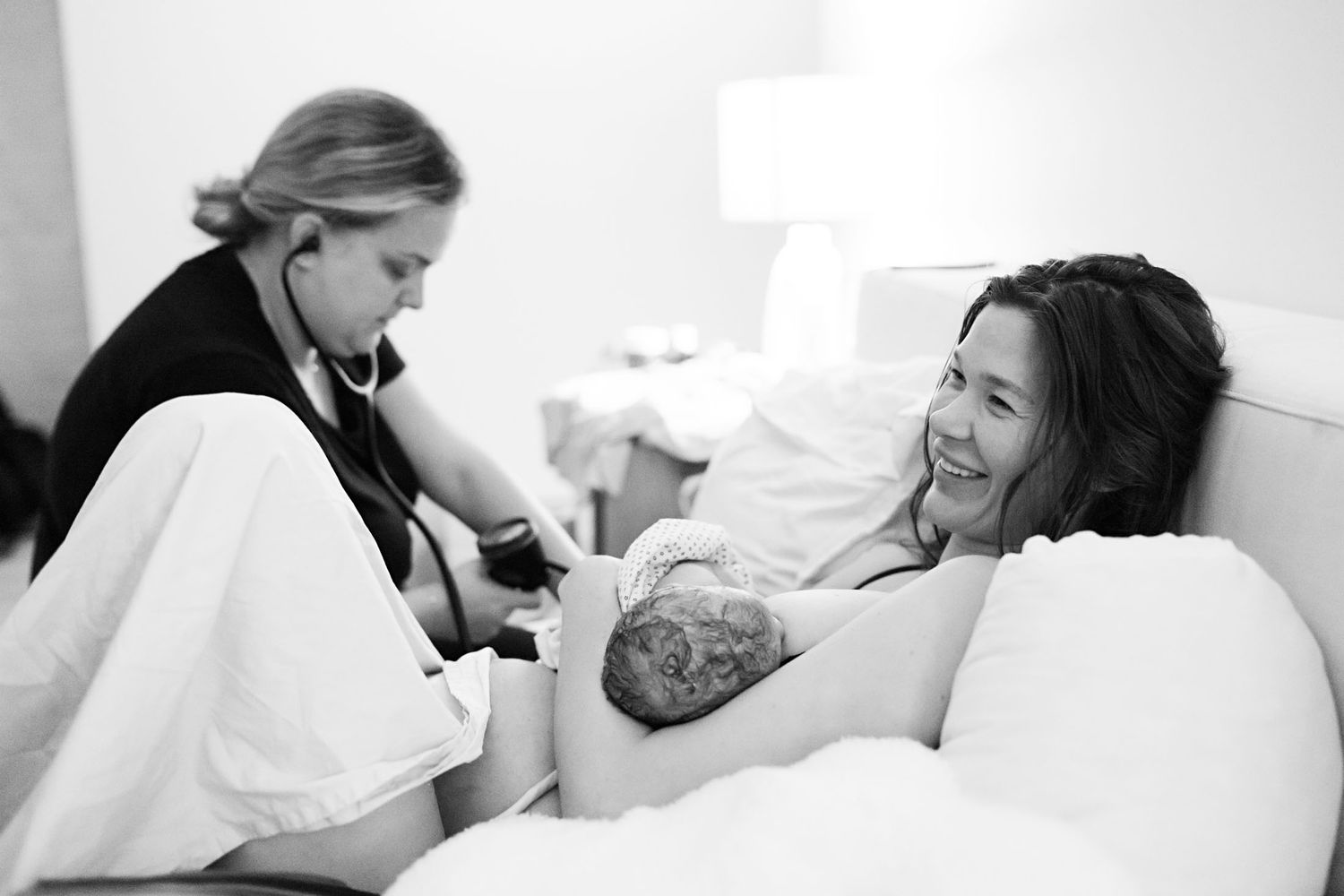 Midwife examines newborn while happy mother holds baby in bed after home birth.