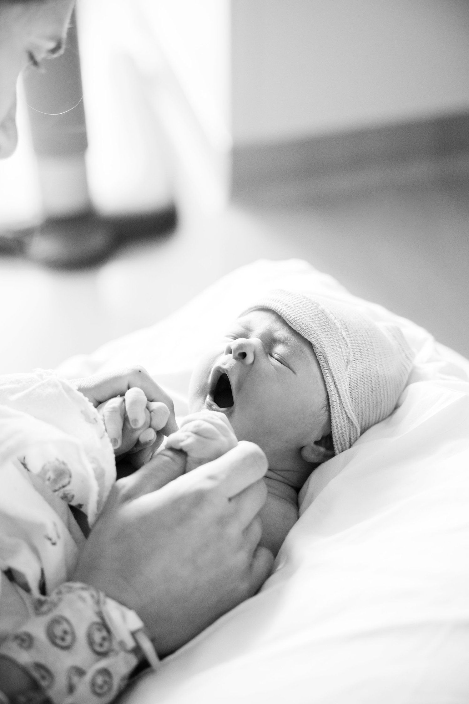 Black and white photo of a newborn baby yawning while being held in a hospital, wearing a knit hat.