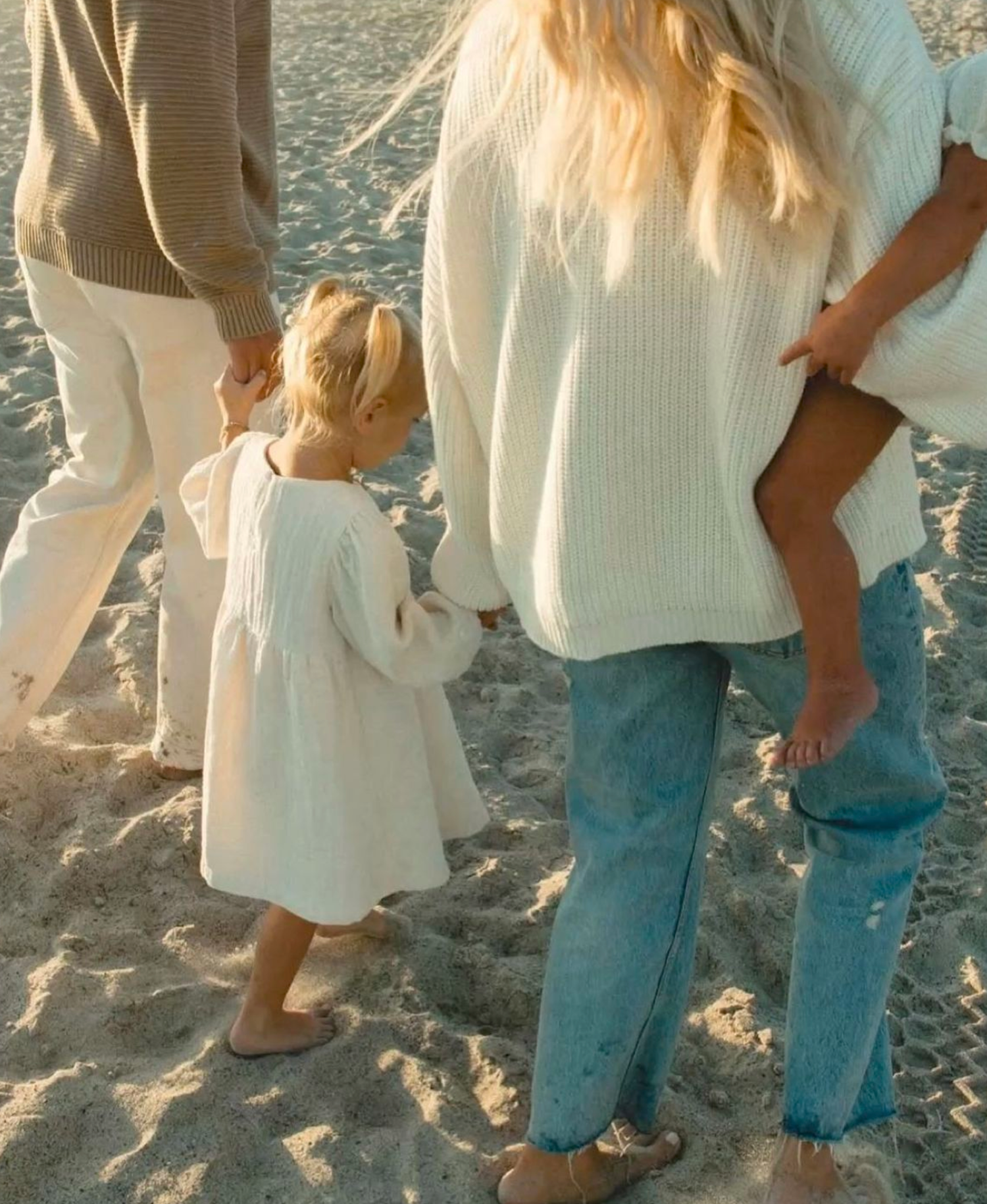 Family walking on sandy beach, toddler in white dress holds parents hands, woman in cream sweater and blue jeans.