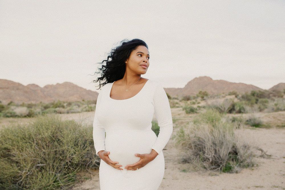 Pregnant woman in white dress cradling baby bump in desert landscape with mountains in background.